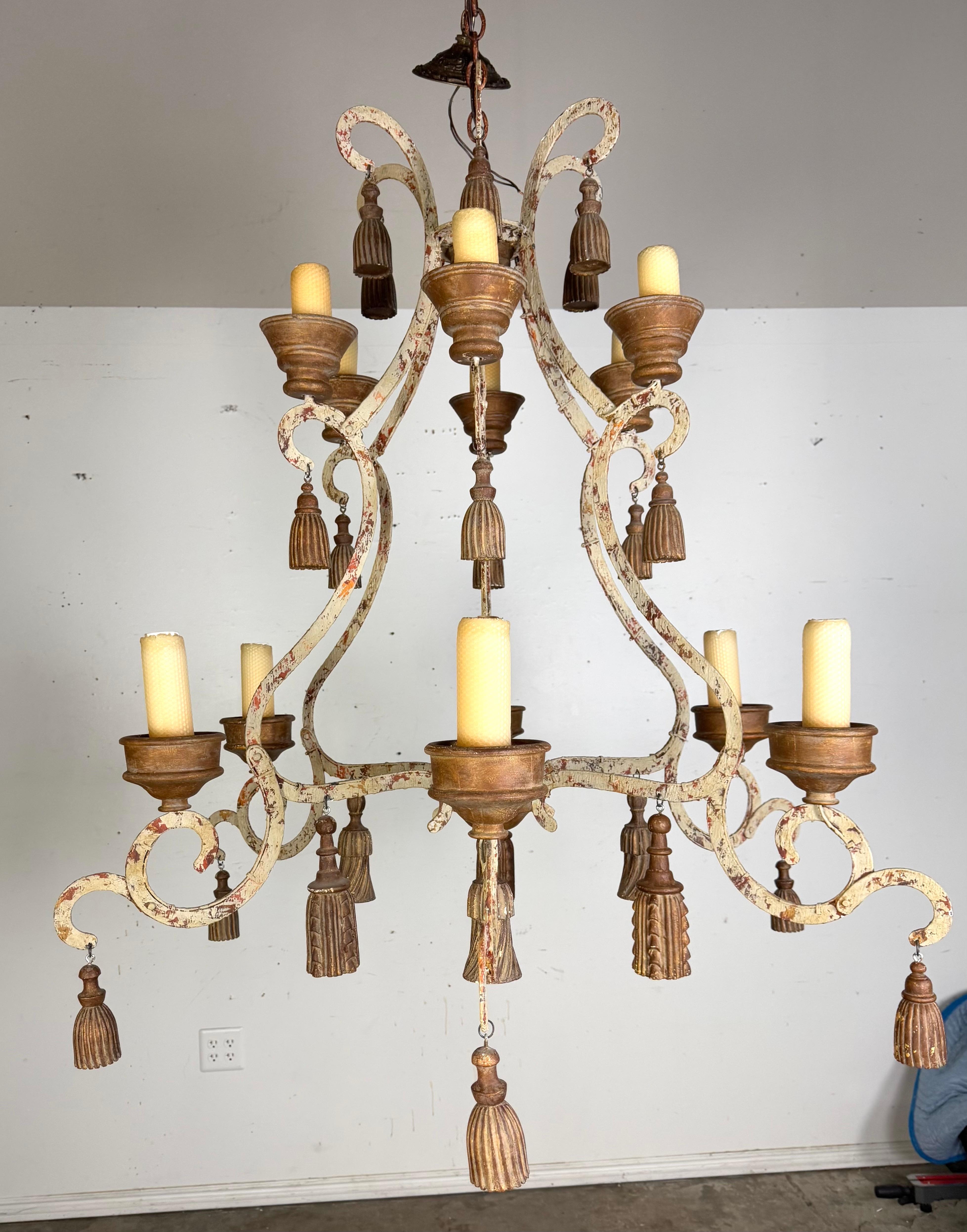 Stile provinciale francese  lampadario a due livelli in ferro battuto ornato da nappe dorate.  La struttura forgiata a mano presenta un'intricata disposizione di bracci che scorrono graziosamente, verniciati con una finitura anticata.  Ogni braccio