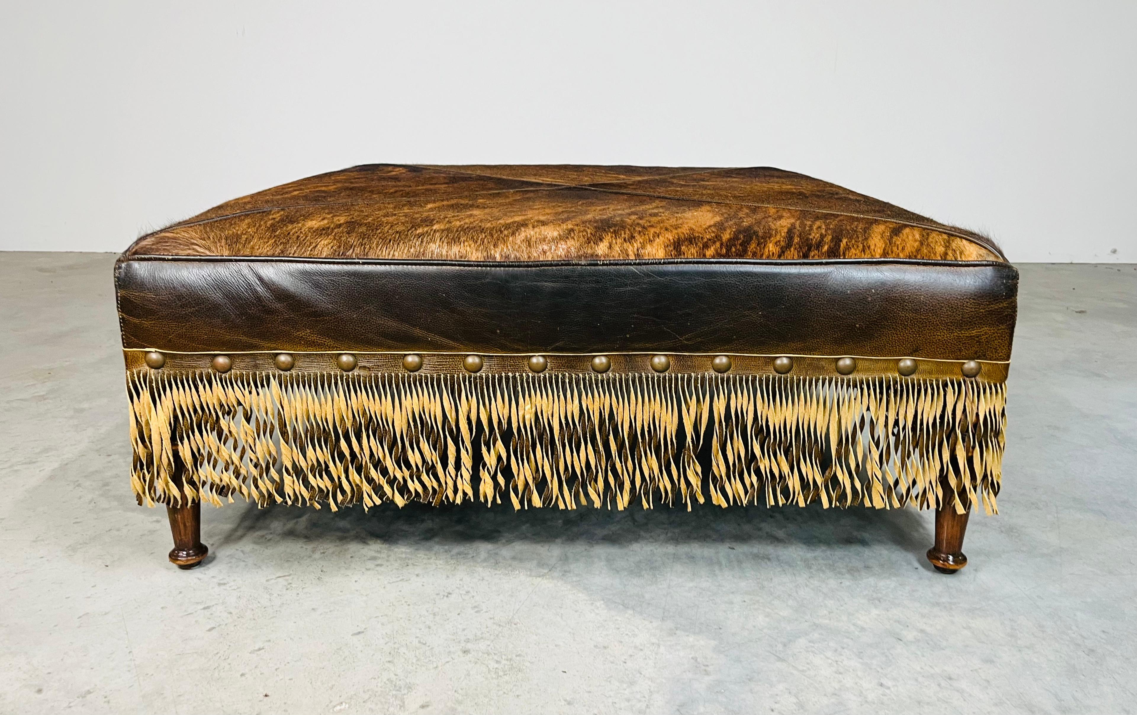 An elegant yet bold cocktail ottoman bench in the French Provincial style having brilliant cowhide top, aniline leather sides with leather fringe surround over turned mahogany legs. Manufactured by Old Hickory Tannery in 2006. Features full aniline
