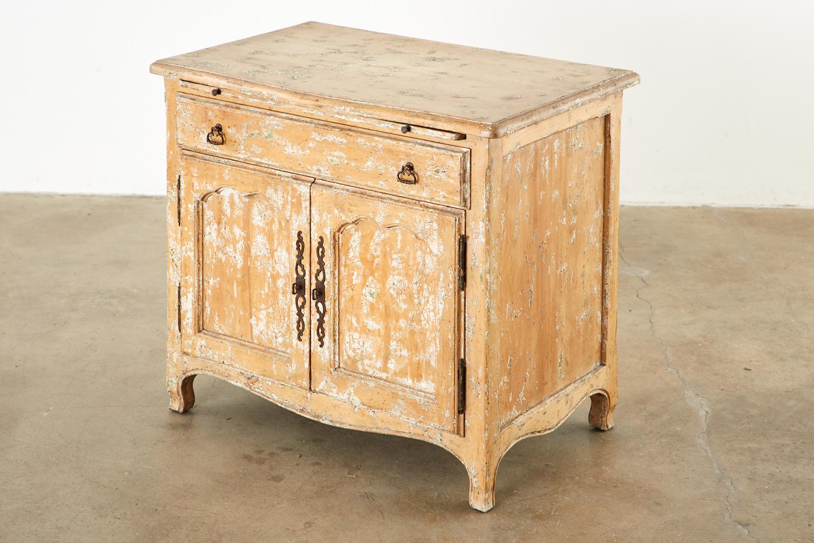 Intentionally aged French provincial style nightstand cabinet. Featuring a pull-out tray or writing table. Beautifully crafted with a pickled lacquer finish having paint remnants to mimic age. Fronted by a large storage drawer and two doors below