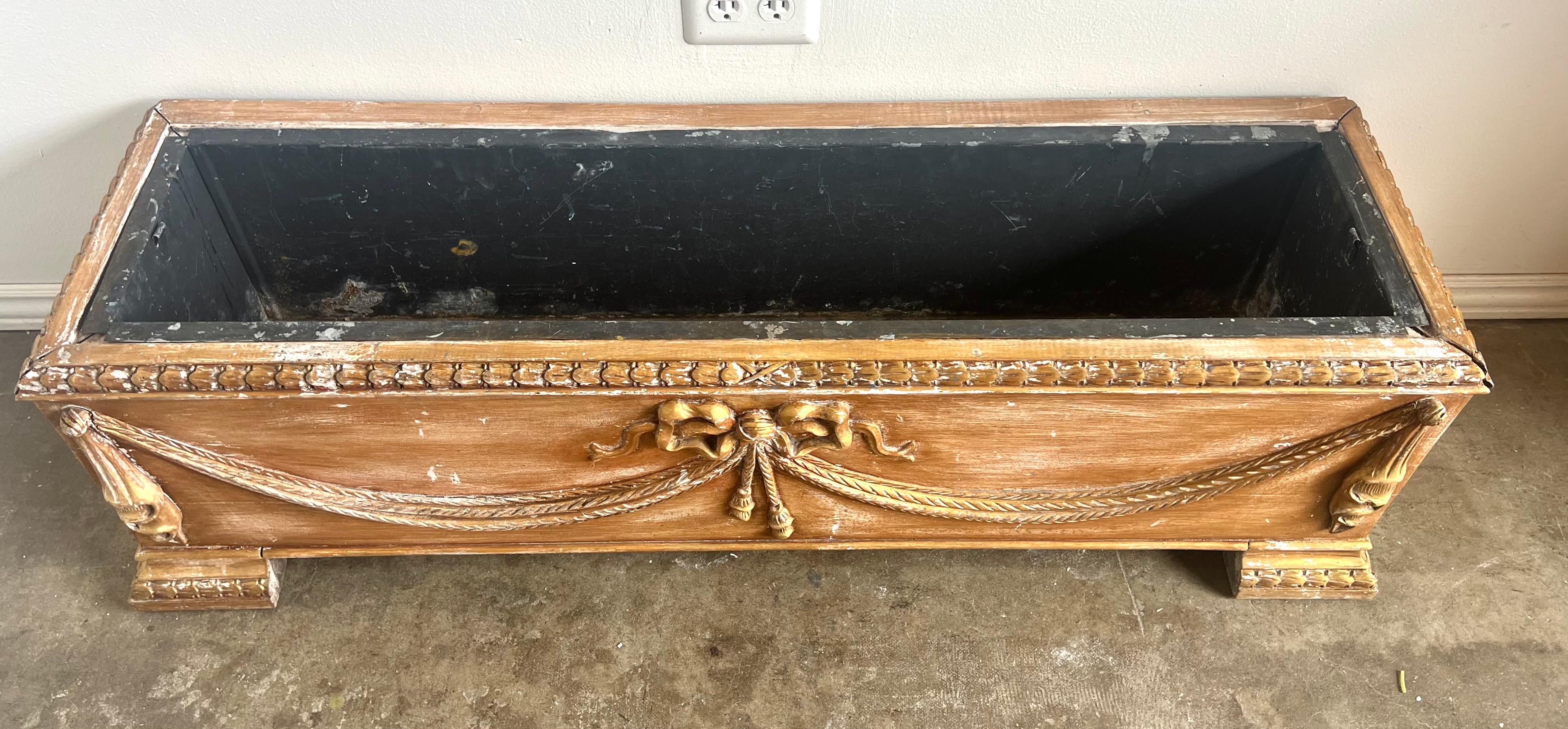 French elongated, rectangular planter with ornate decorative elements.  
The planter is crafted with a wooden frame that features a distressed finish, enhancing its vintage appeal.  The front and sides are embellished with gilt details including a