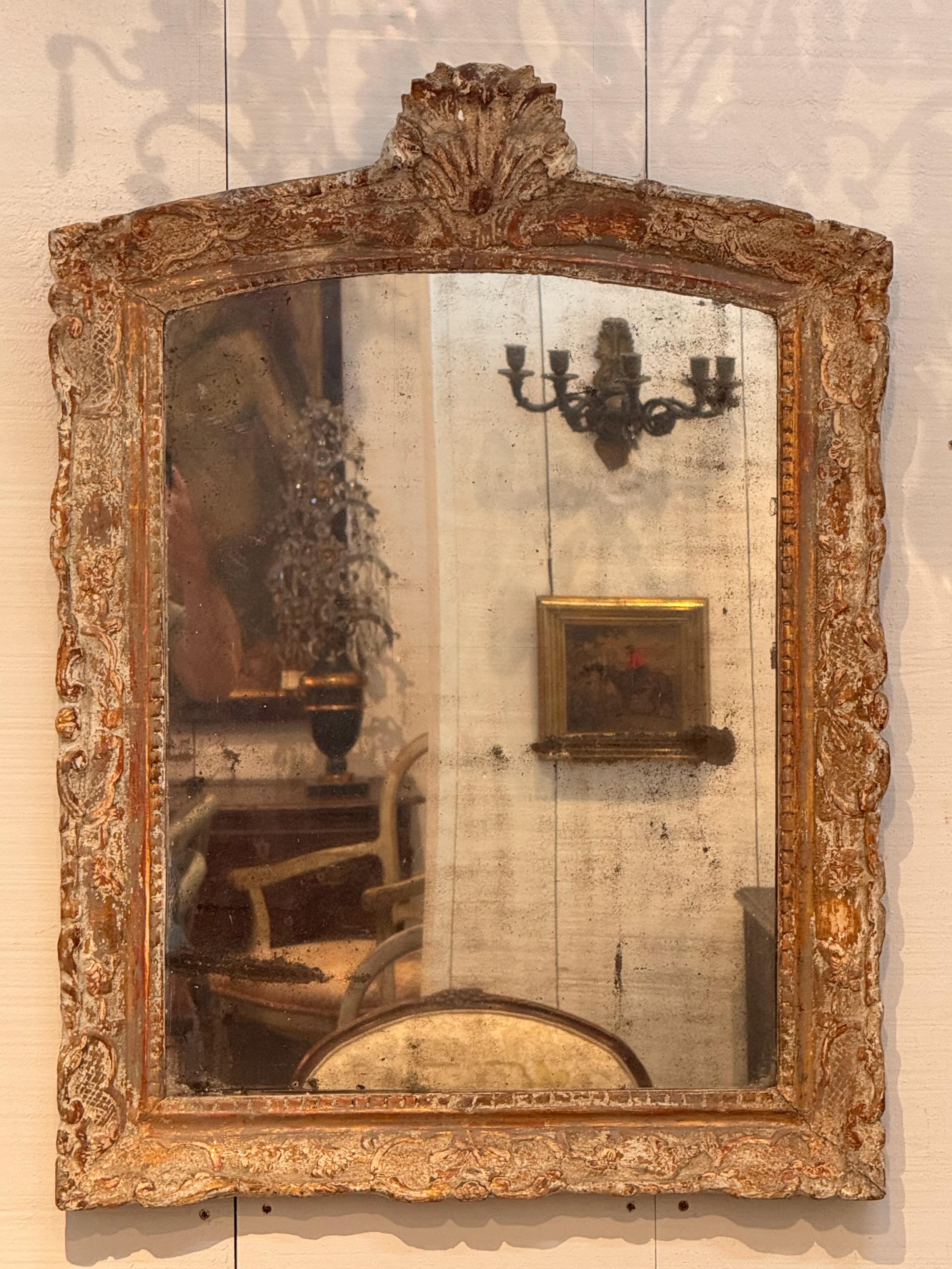 French Regence-style mirror featuring a richly carved and giltwood frame with an aged patina. The arched top is crowned with a dramatic shell motif flanked by scrolling foliage, while the rest of the frame displays intricate relief carving. The