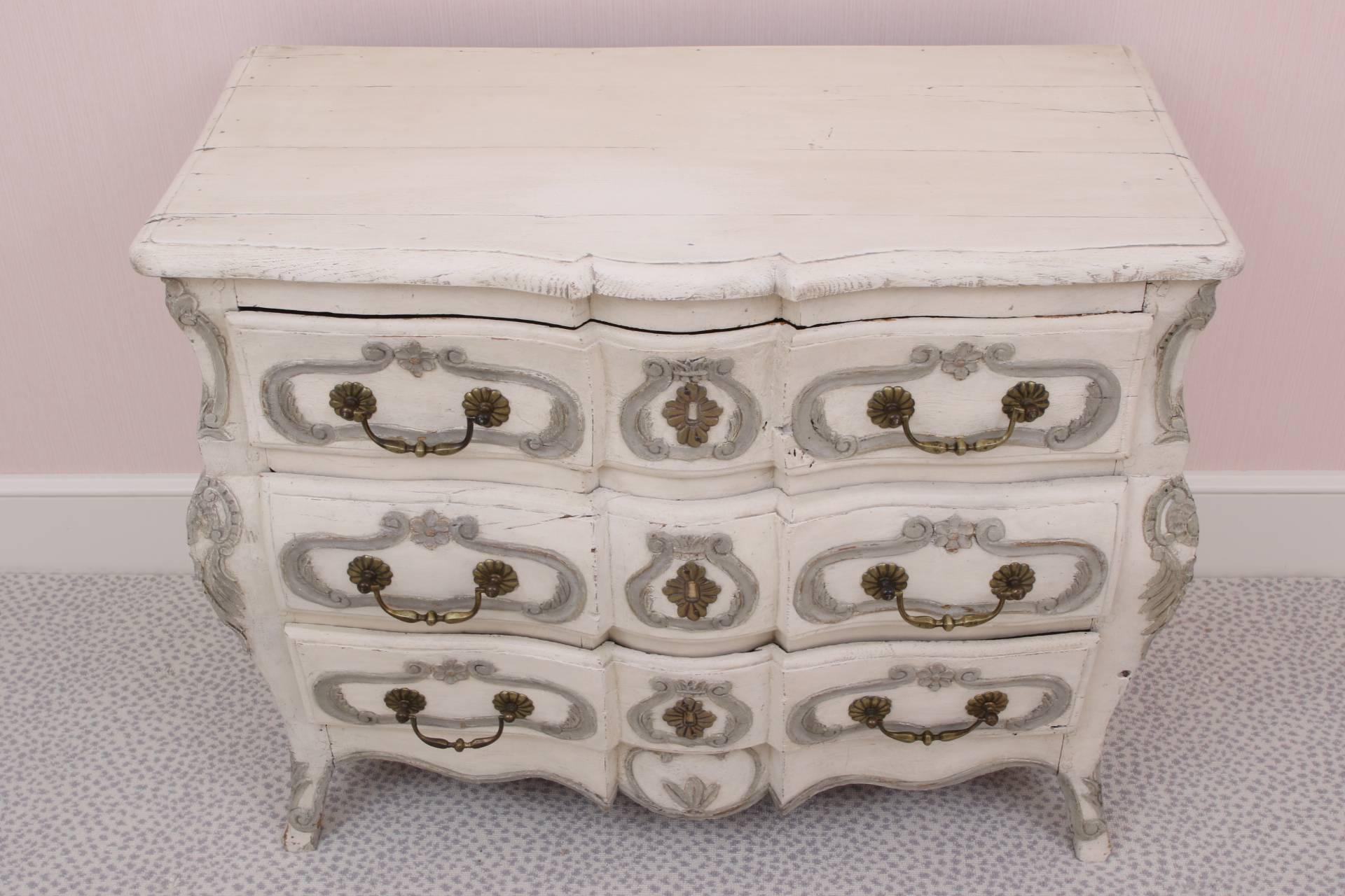 In cream paint with carved decoration in pale grey. A plank top and recessed shaped sides, three drawers with grey scrolled motifs, the centre ones with faux escutcheons, and fine bronze handle pulls. The side frames with grey cartouches and leaf