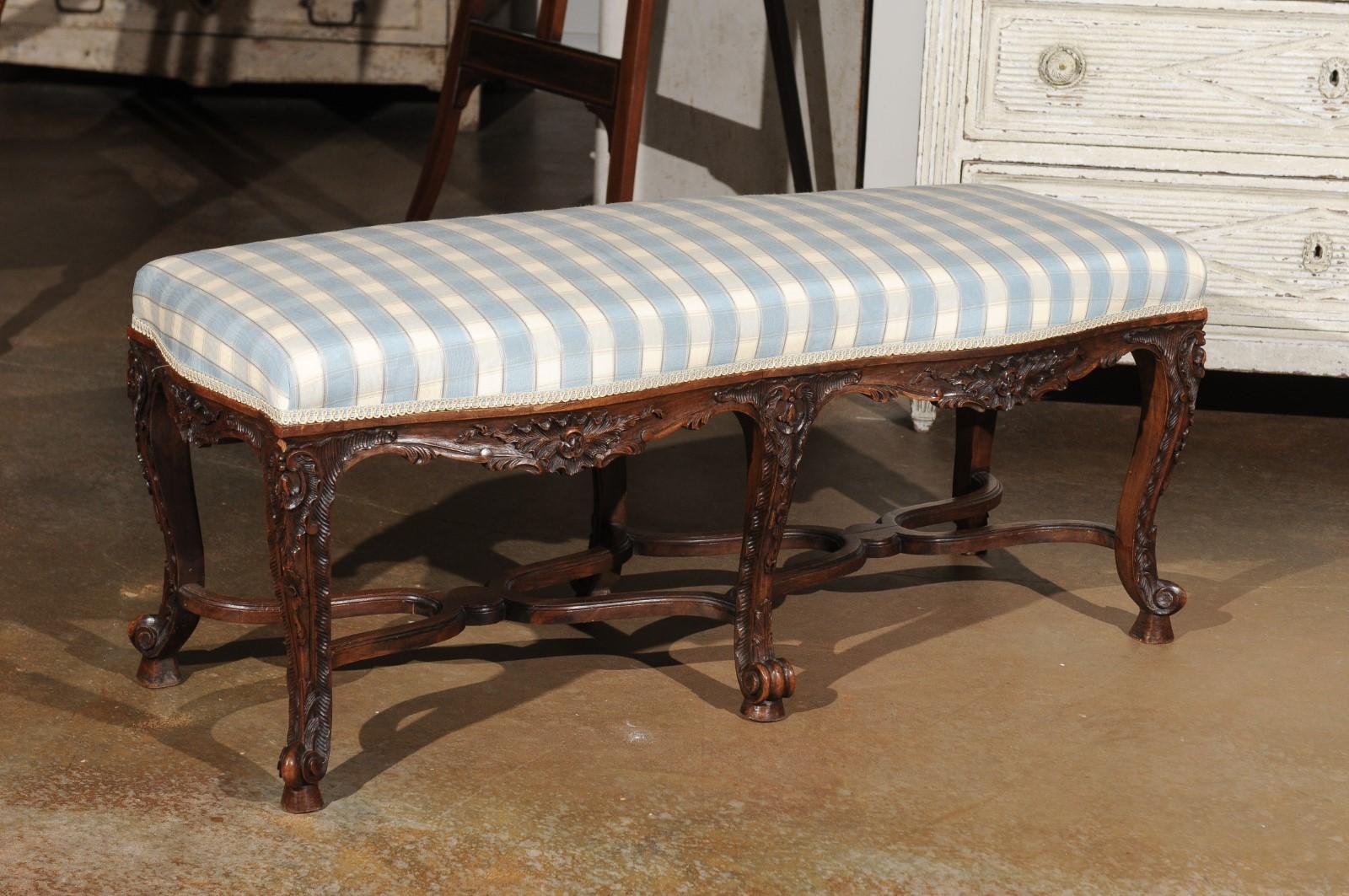 A French Regence style carved wooden bench from the 19th century, with X-form cross stretcher, foliage décor and new checkered upholstery. This exquisite French backless bench features a rectangular seat recovered with a soft blue and white checkred