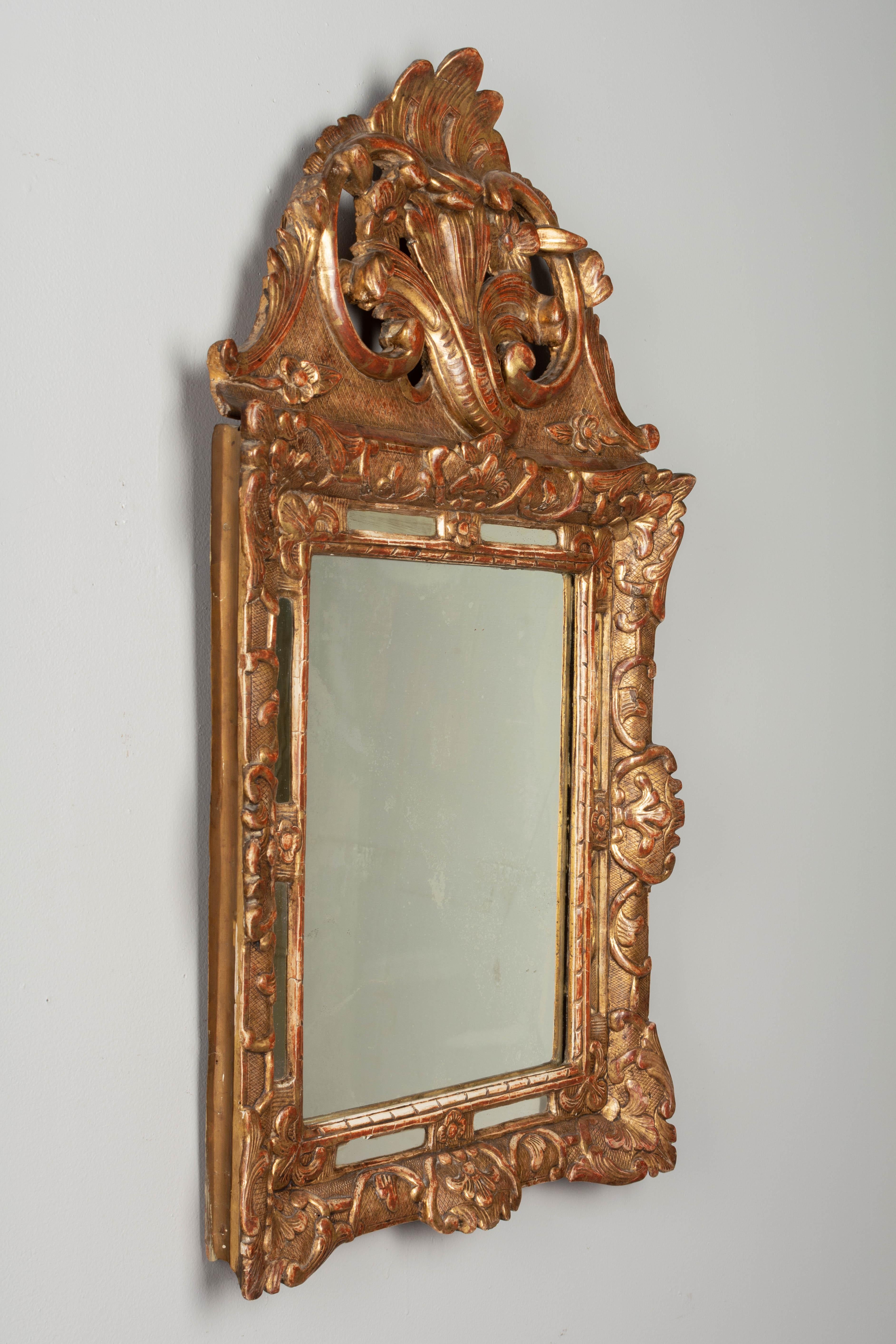 An early 19th century French Regence style giltwood parclose mirror with large boldly carved sculptural crest and elaborate floral and leaf form corner decorations. Warm gilt finish with red clay undertone. Original mirror with old silvering. Minor