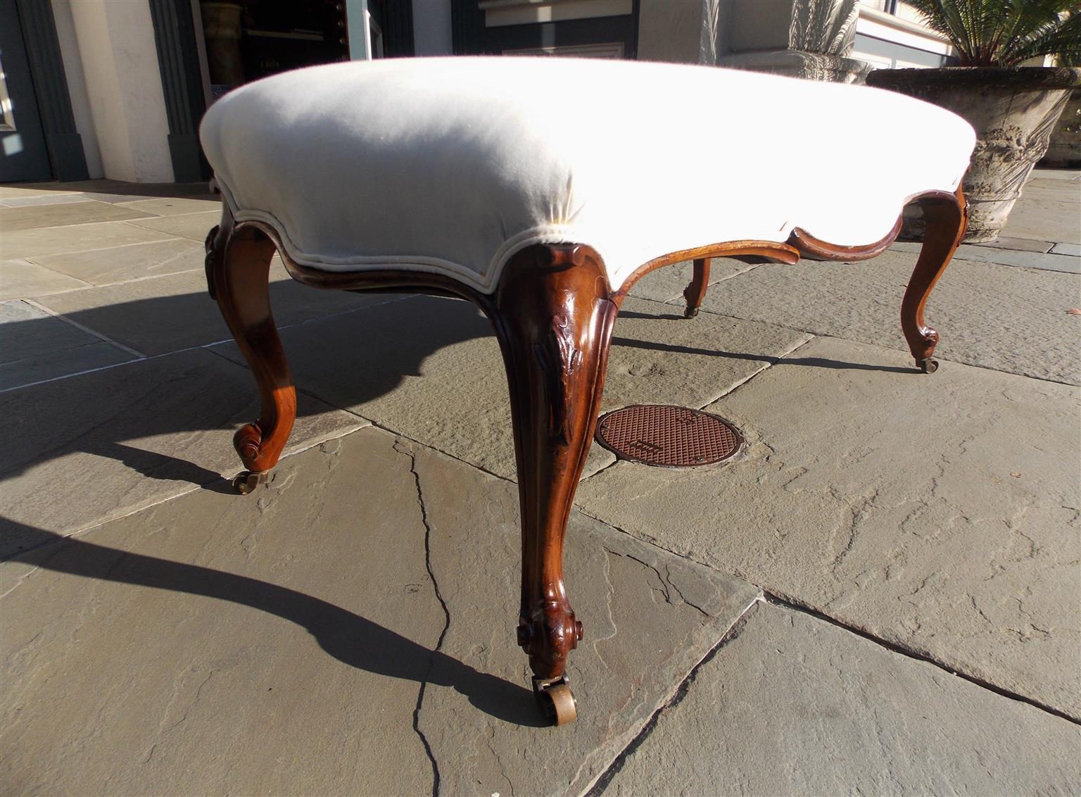 French Regency mahogany upholstered bench with serpentine skirt, foliage carved knee, and terminating on the original brass casters. Bench is covered in white muslin with horse hair and cotton padding, Early 19th century.