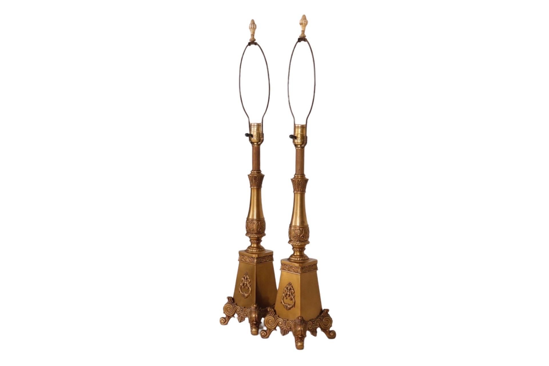 A pair of French Regency style table lamps made of brass. A short fluted column sits above a baluster shaped central column decorated with branches of olive leaves around the neck, and scrolls with acanthus leaves around the bottom. Below, a plinth