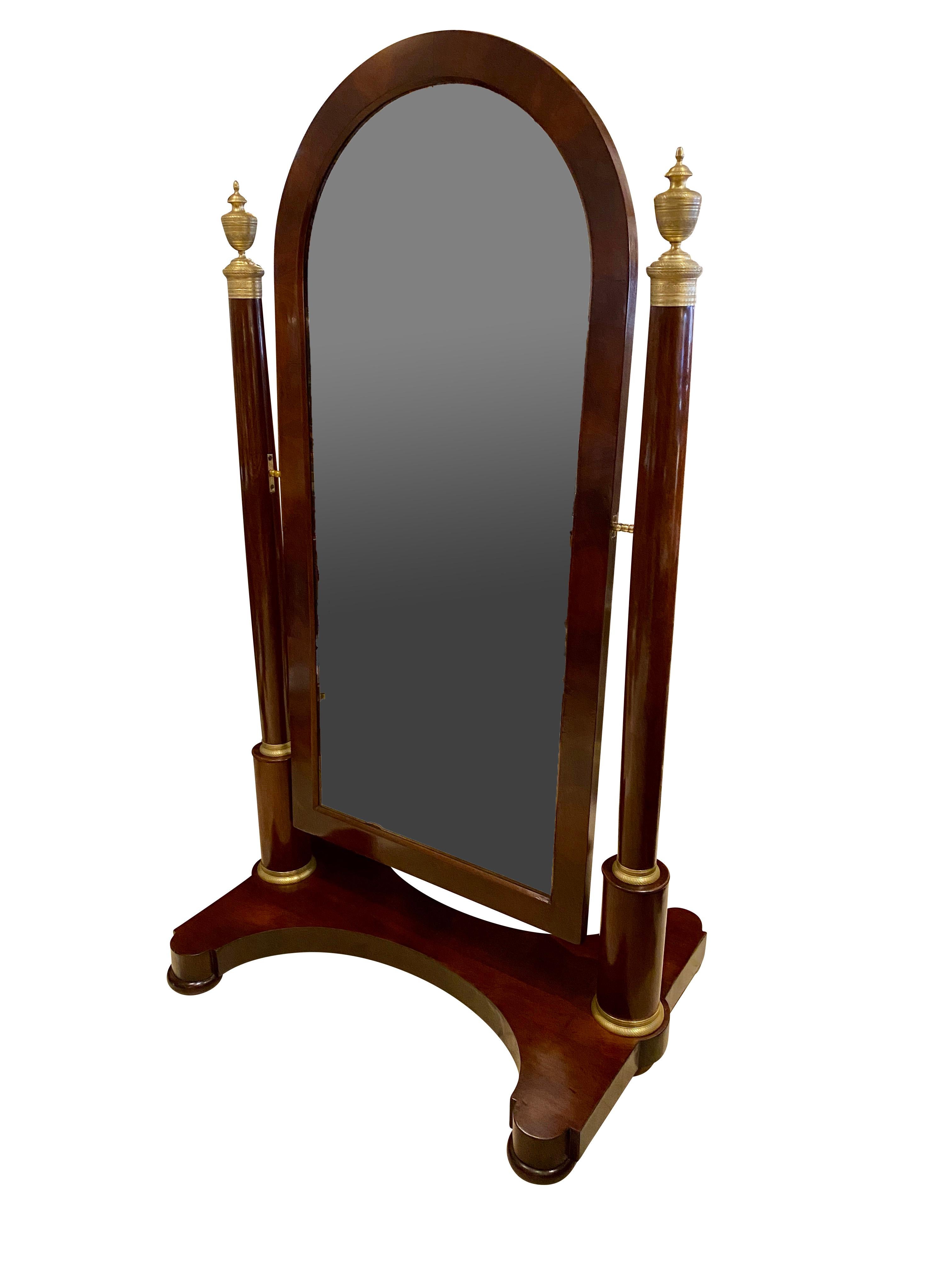 With an arched mirror plate set in a conforming wood frame that swings, all attached to a pair of cylindrical posts each with engine turned urn form finials, the double U-form base with disc form feet.