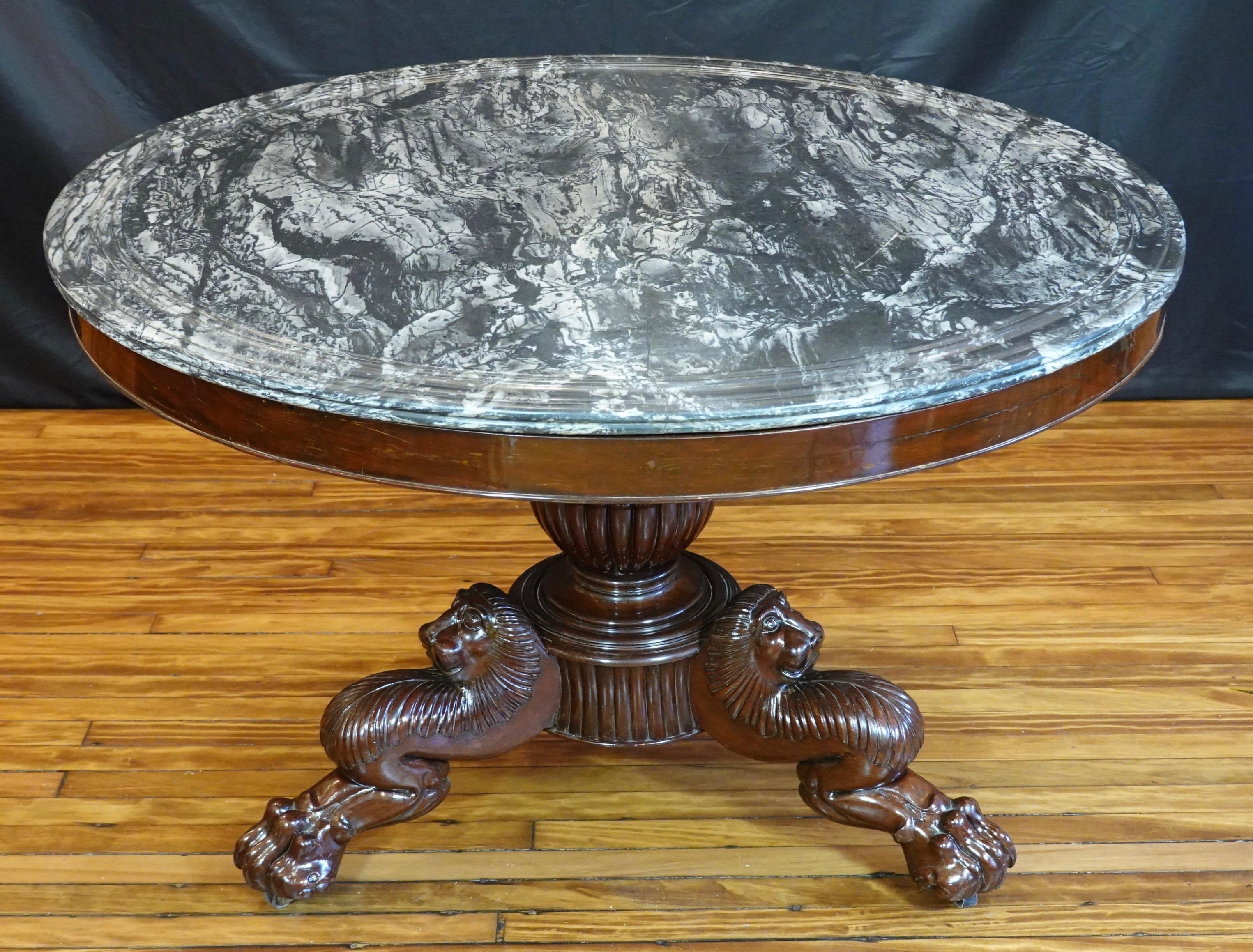 Large French Restoration period (circa 1815-1830) mahogany center table with variegated black and white marble top. The base stands on a central lobed pedestal with three legs with impressive carved lion's heads terminating in paw feet with casters.