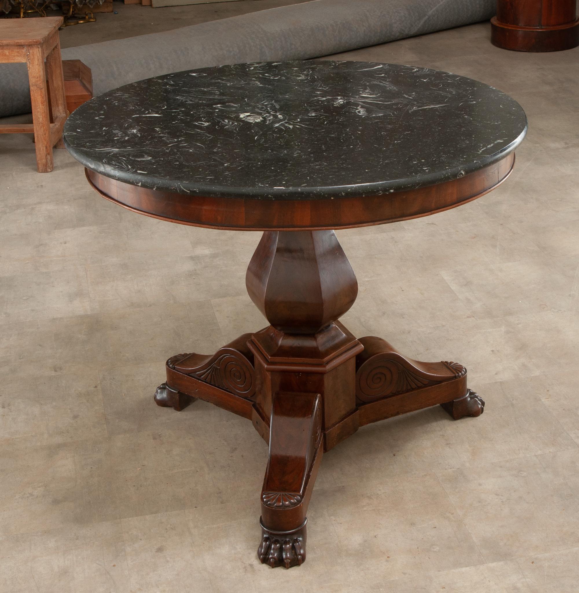 A French marble top gueridon from the Restauration period. This round table is topped with a black fossil marble with a carved lip. Below is a detailed mahogany pedestal base with scroll legs ending in paw feet. Cleaned and polished with a French
