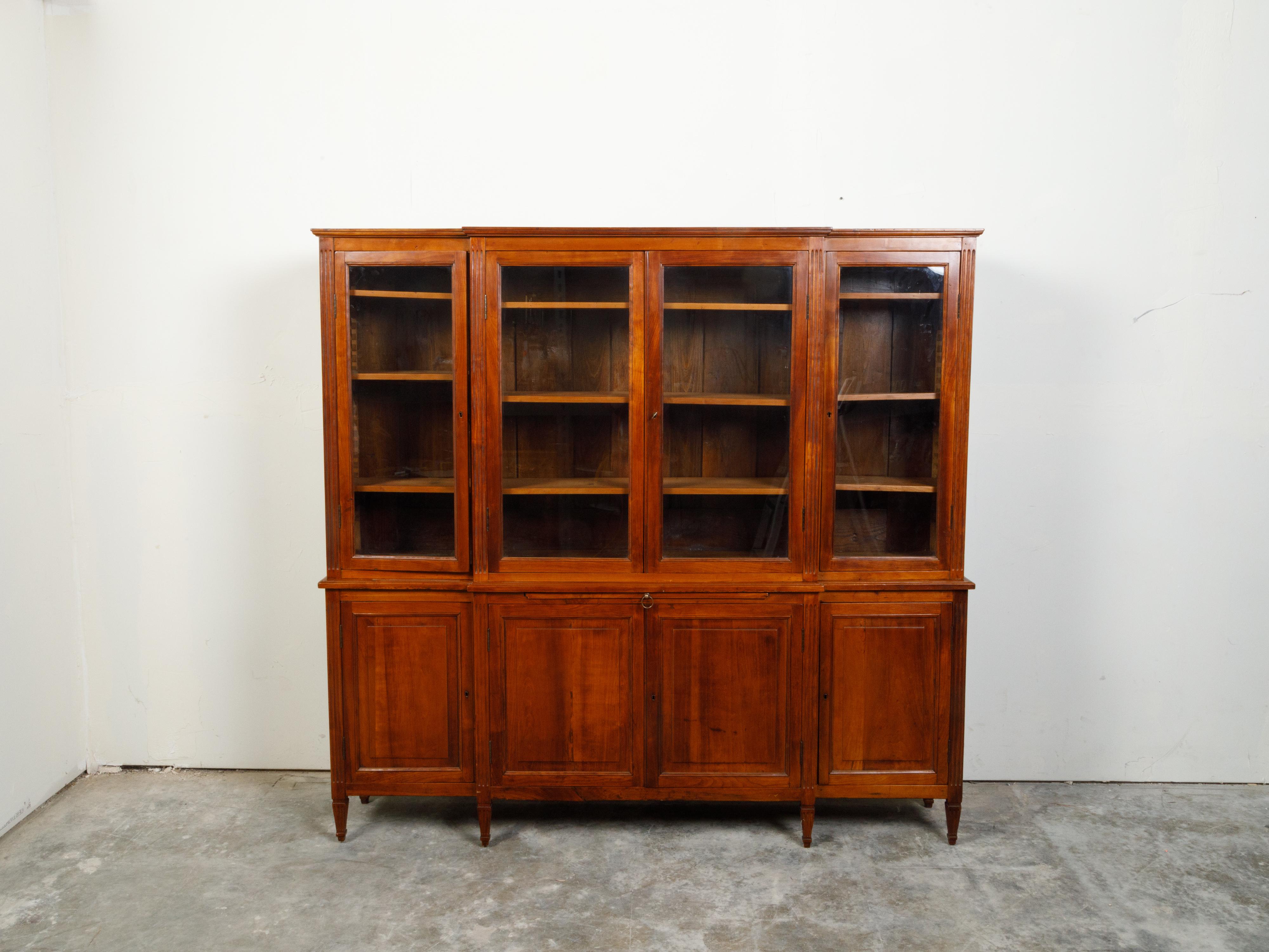A French Restauration period walnut breakfront bookcase from the early 19th century, with glass doors and fluted accents. Created in France during the first quarter of the 19th century, this breakfront bookcase features four glass doors in its upper