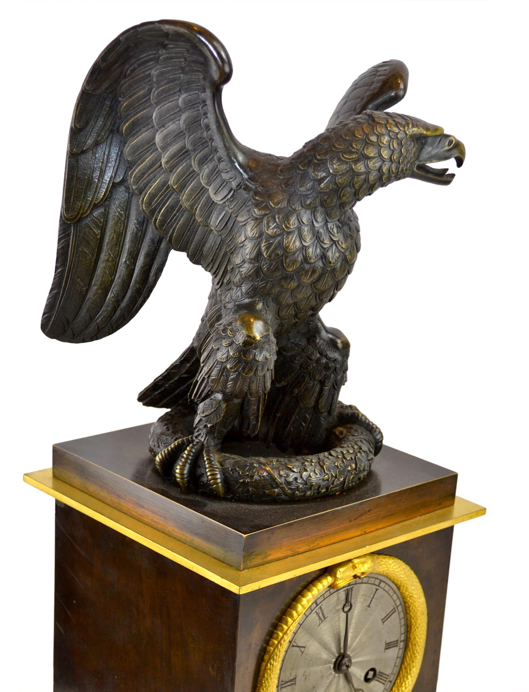 A French restauration period mantle clock having a large patinated bronze eagle with outstretched wings standing on a wreath on top of the clock.

The stepped square patinated bronze base is richly decorated in gilded bronze; the gilded snake bezel