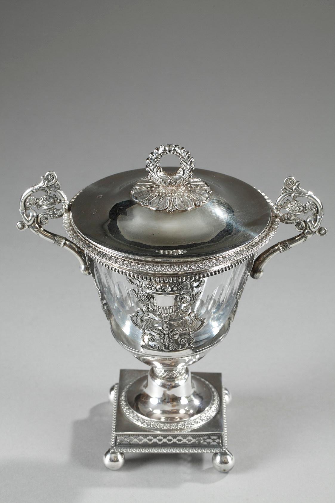 Lightly tapered candy dish in silver and crystal. The rim is ornamented with an openwork geometric frieze, and the paunch features a sculpted silver balustrade vase and two birds standing facing each other on either side of the vase. The birds are