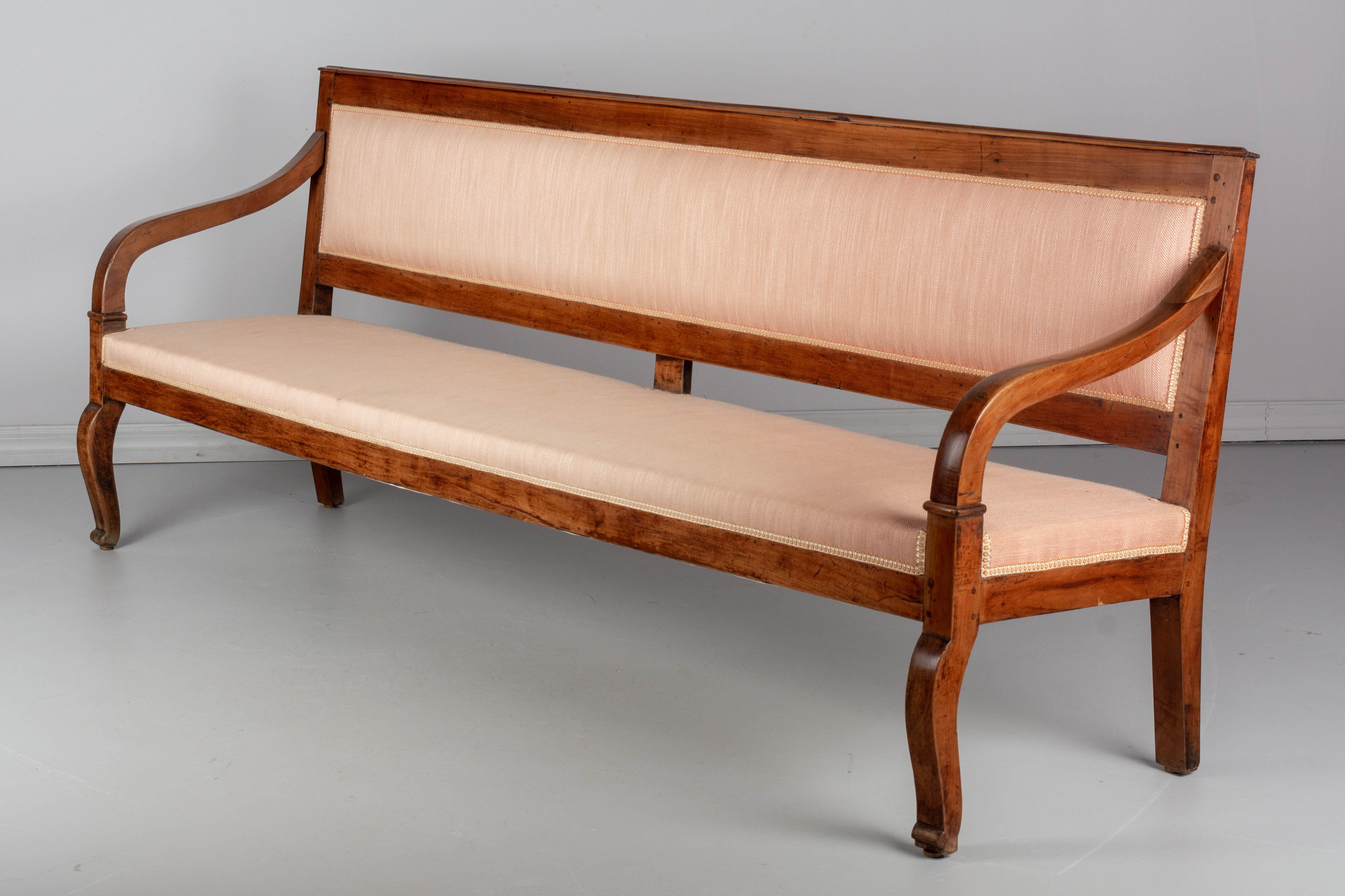 A 19th century French Restauration style bench or banquette, made of solid cherrywood. Sturdy and well-crafted using pegged construction. Old upholstery, as found, is serviceable. An unusually long size, perfect for an entry hall. One of the arms