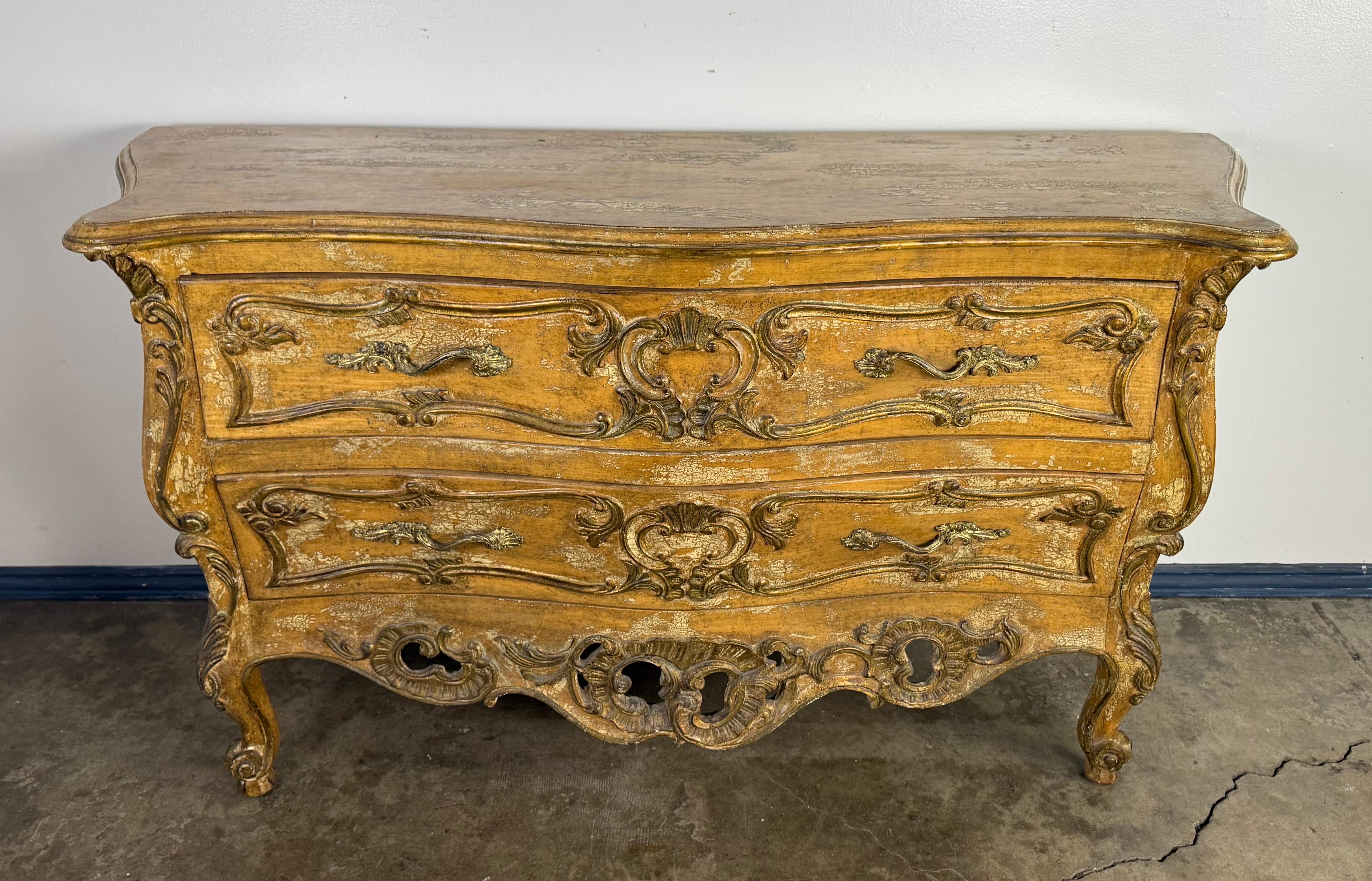 An elegant French Rococo–style commode with beautifully curvaceous serpentine form, carved in richly detailed relief and finished in an aged ochre and gilt patina. The two spacious drawers feature finely scrolled foliate motifs and gilt accents,