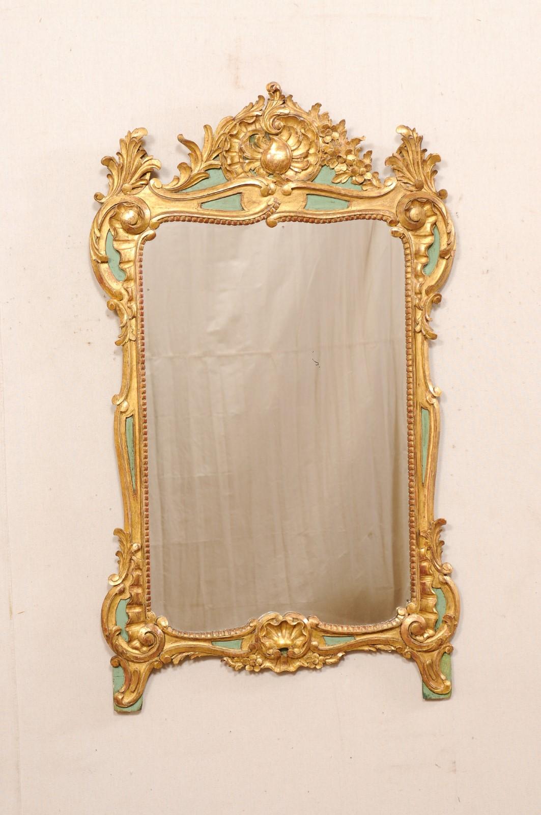 A French rococo style carved and gilt/painted wood mirror from the early 20th century. This antique mirror from France features a rectangular- shaped mirror with shapely surround in a gilt wood with elaborately carved rinceaux with shell at top