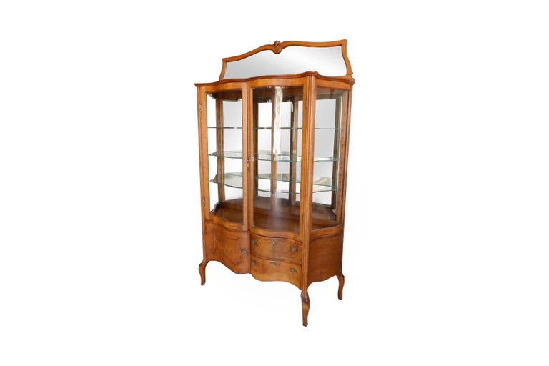 A French Rococo style china cabinet made of oak with a mirrored back. An upper vitrine with curved glass doors houses three serpentine glass shelves and is surmounted with a beveled mirror. Below to the right are two drawers and to the left is a