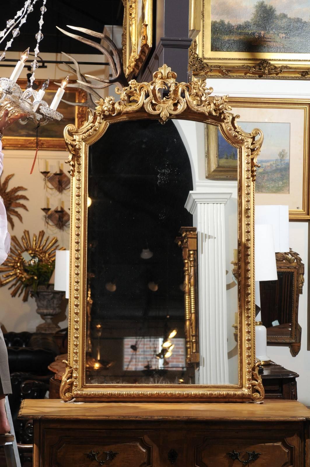 A French Rococo style giltwood mirror with carved crest from the 19th century. This vertical French Rococo style mirror features a trapezoidal frame, adorned with delicate beads, and egg motifs set inside a guilloche style frieze. Two acanthus