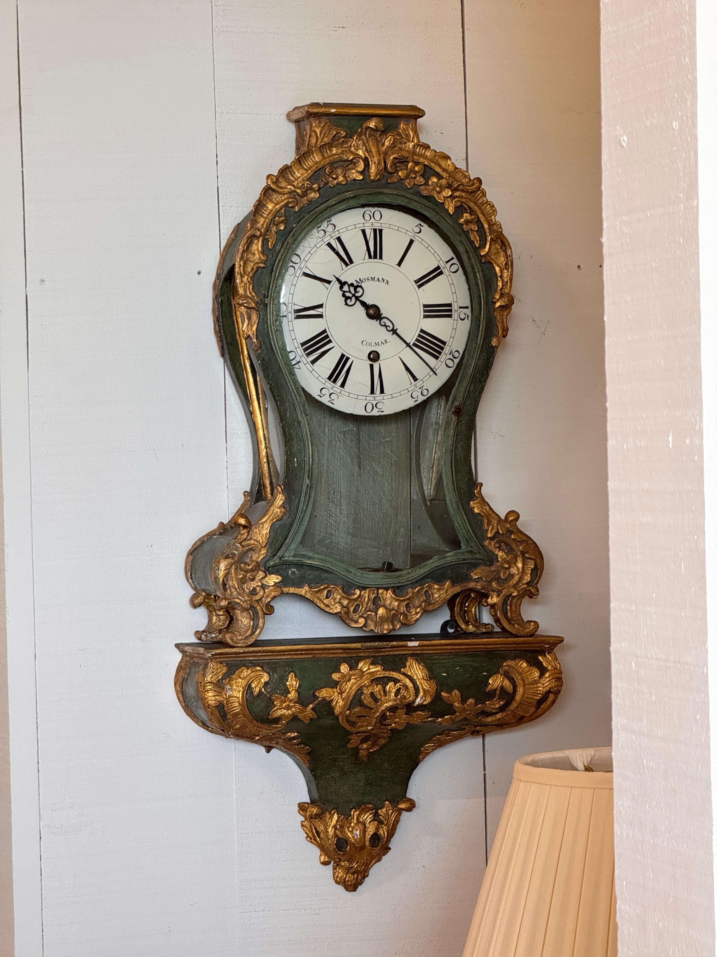 Decorative French mantel clock with a scrolled and gilded wood case in the Rococo style. The painted green finish is accented with gilt floral and foliate details. The enamel dial is signed “Mosmann, Colmar” and features Roman numerals with a glass