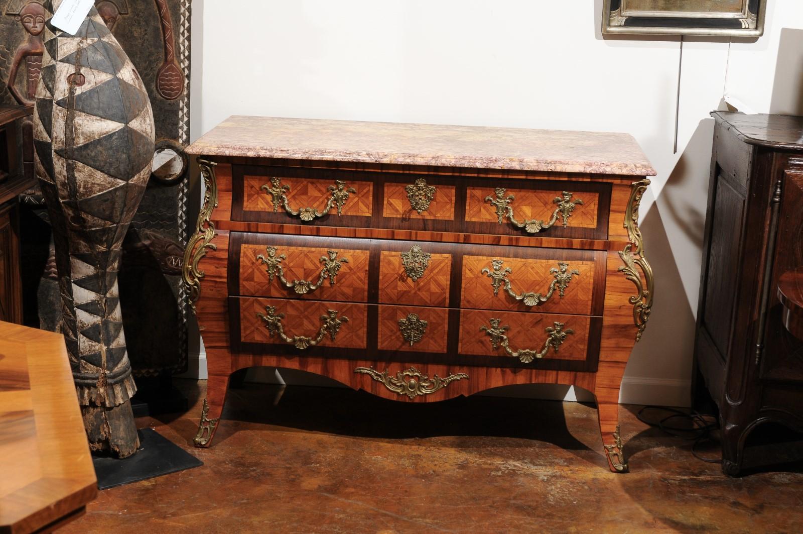 A French Rococo style bombé marquetry inlaid commode from the early 20th century, with marble top and bronze hardware. This exquisite French Rococo style three-drawer commode features a rectangular variegated marble top with bevelled edges, sitting