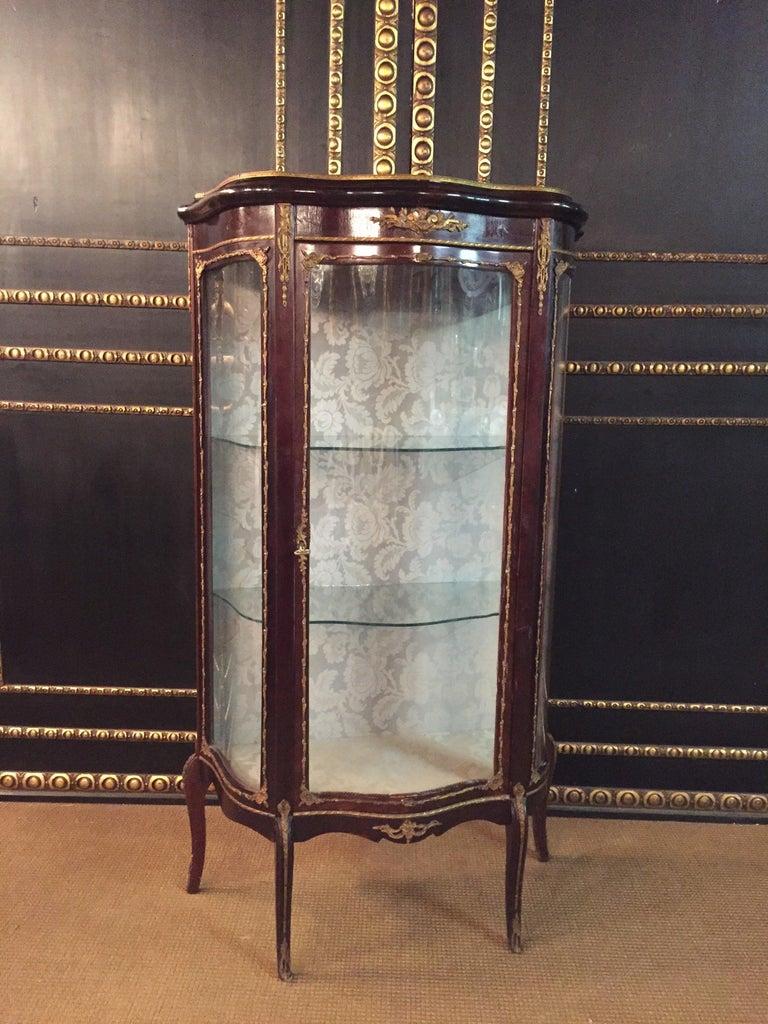Delicate French vitrine rosewood on solid wood. High-rise, one-door, cambered and three-sided glazed body. On curly feet scalloped out. Three-sided glazed vitrine. Slightly protruding top plate with brass gallery. Rich bronze fittings. Backboard