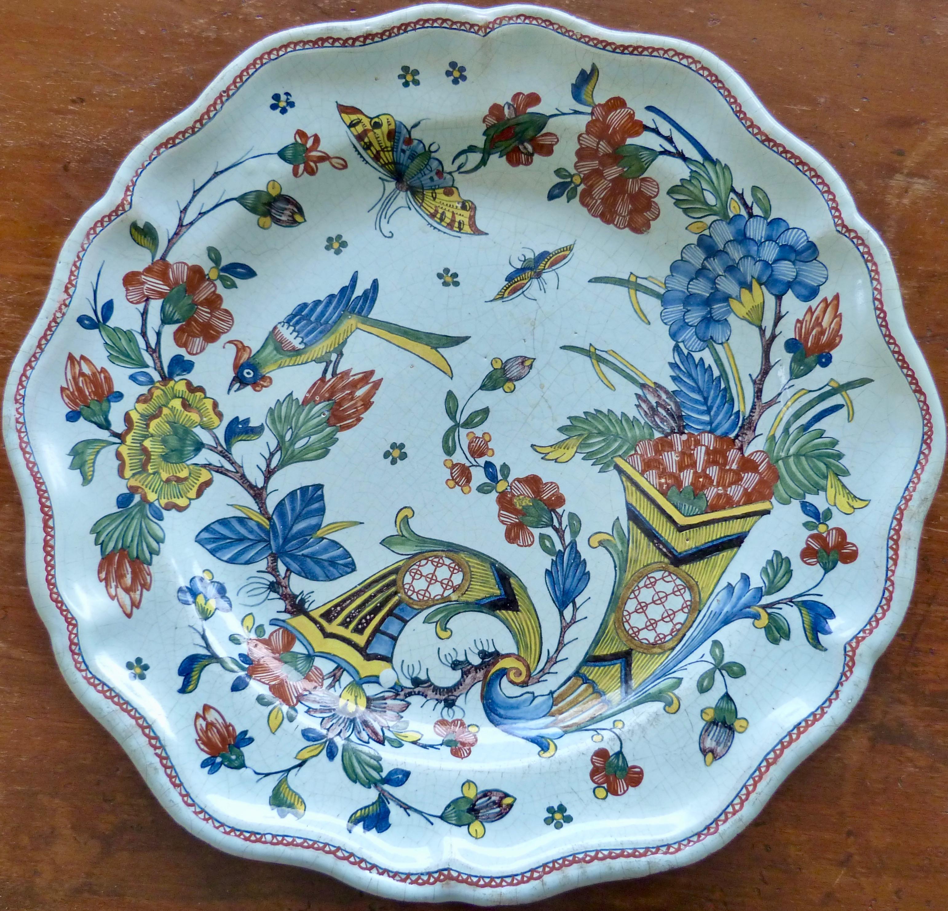 A French Rouen large Faïence charger with scallop-shaped edges. Picturing the sign of prosperity double Cornucopia and with blooming flowers, butterflies and a bird. Decorated in a typical polychrome palette of colors composed of green, blue, iron