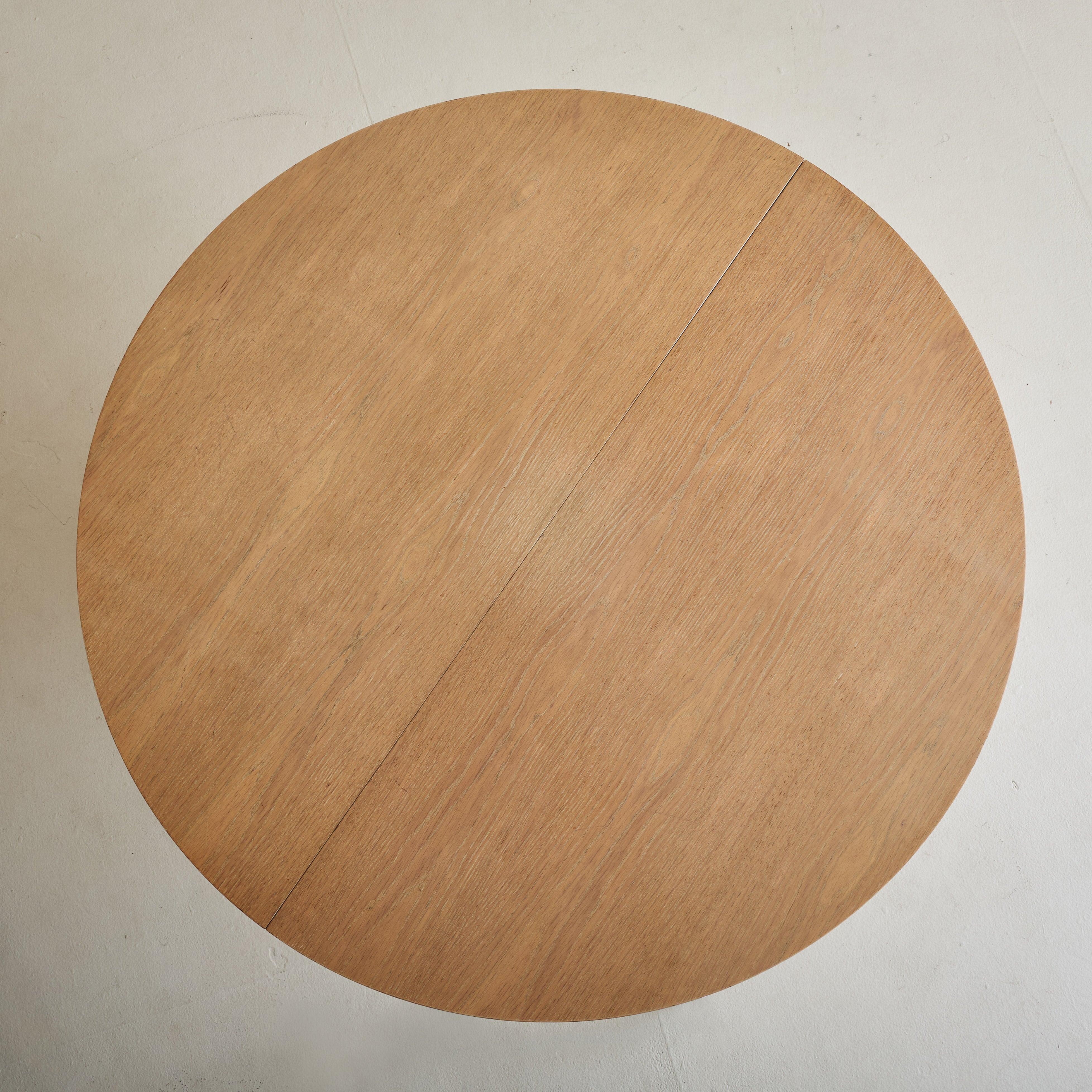 A 1940s French round oak center table with Art Deco influences, characterized by its architectural form. The sculptural pedestal base, composed of two tiers, is accented with geometric brass details. The table is expandable and includes inset