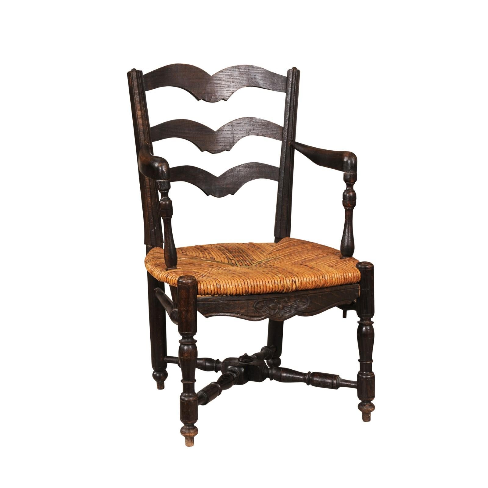 A rustic French dark oak child's chair from the 19th century with carved ladder back, open arms with turned supports, rush seat, carved apron and turned legs with cross stretcher. Introduce a touch of rustic charm to your home with this charming