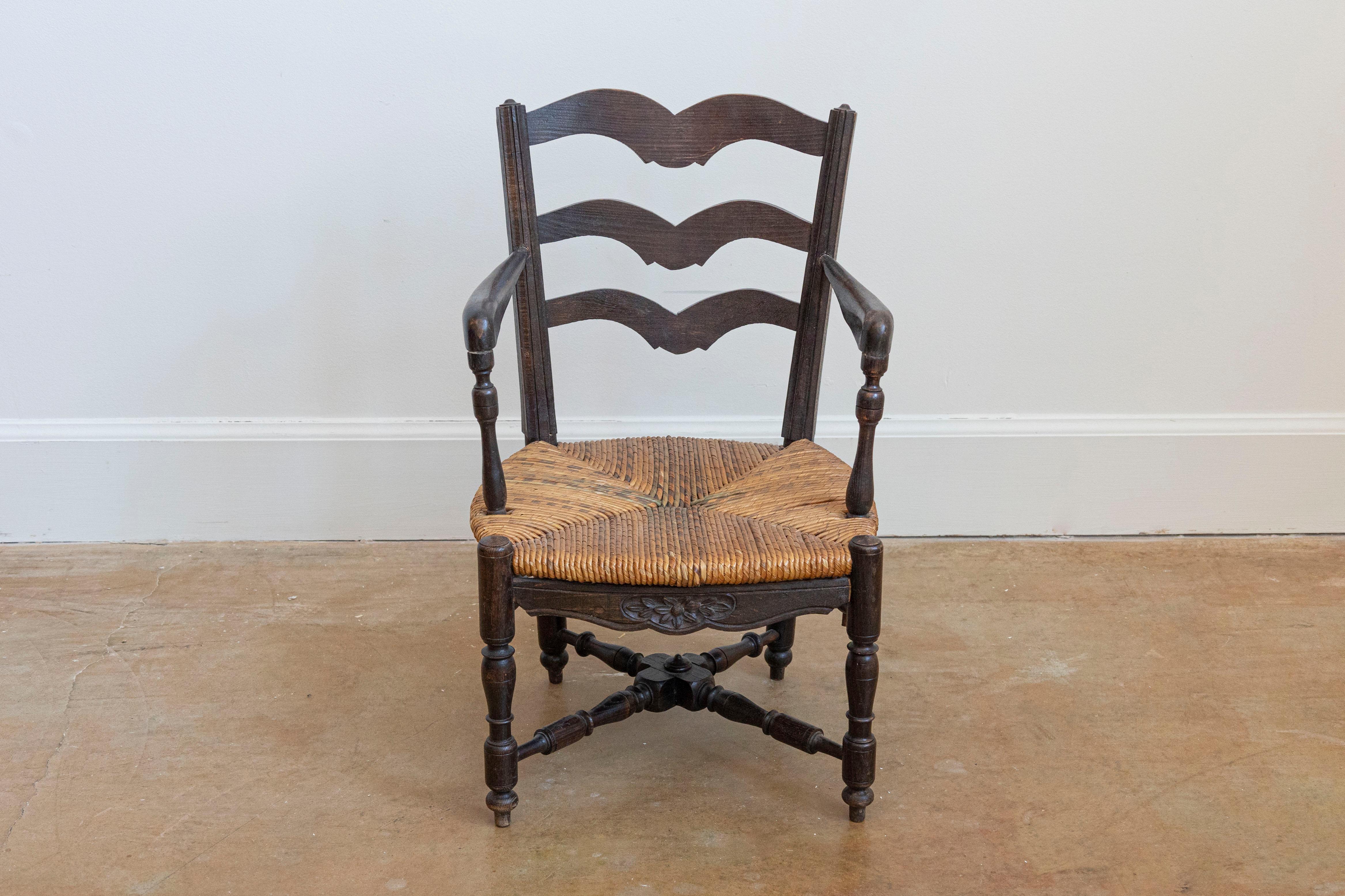 A rustic French dark oak child's chair from the 19th century with carved ladder back, open arms with turned supports, rush seat, carved apron and turned legs with cross stretcher. Introduce a touch of rustic charm to your home with this charming