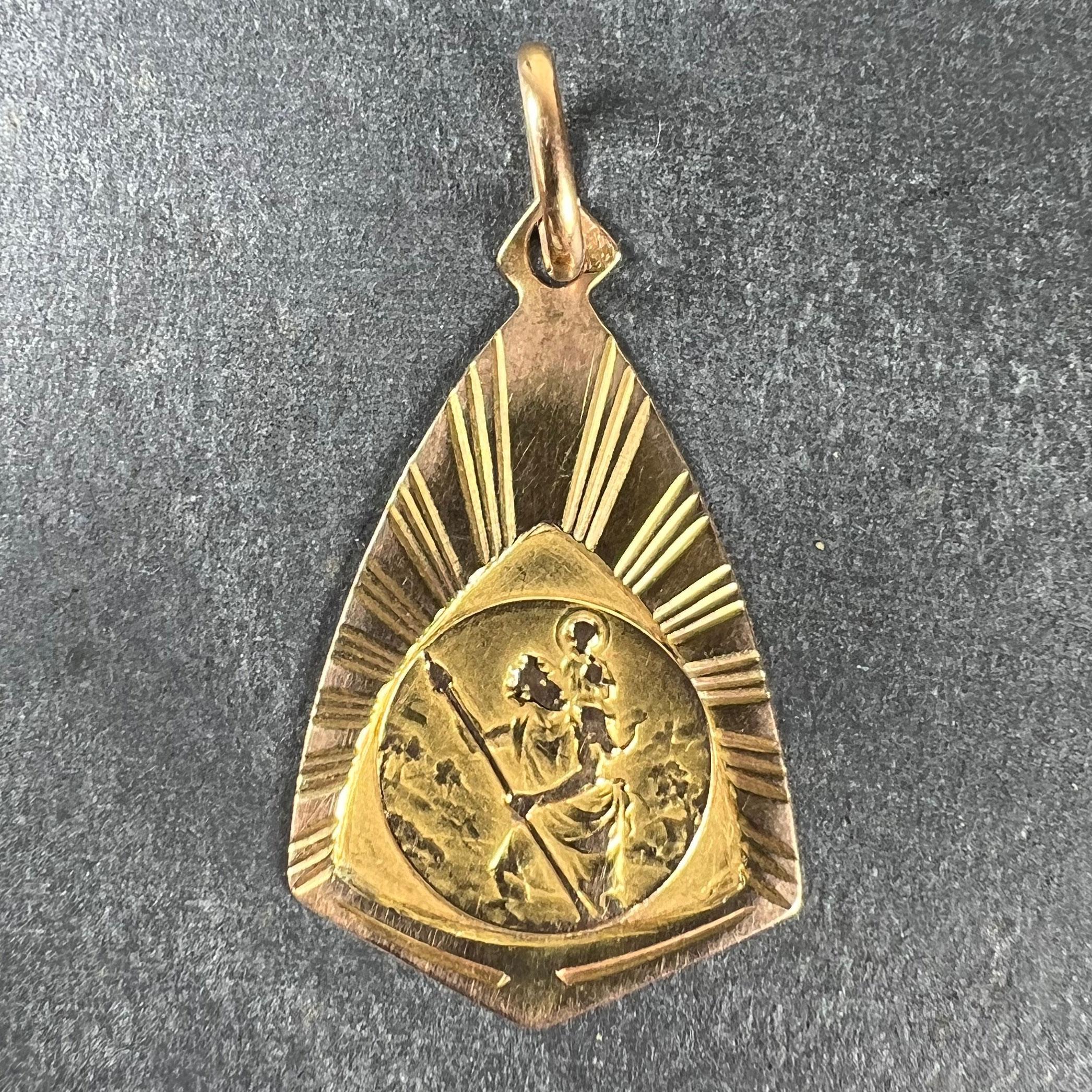 A French 18 karat (18K) yellow gold charm pendant designed as a circular medal depicting St Christopher carrying the infant Christ across the river set within a kite-shape teardrop with radiating engraved lines. Stamped for 18 karat gold with an