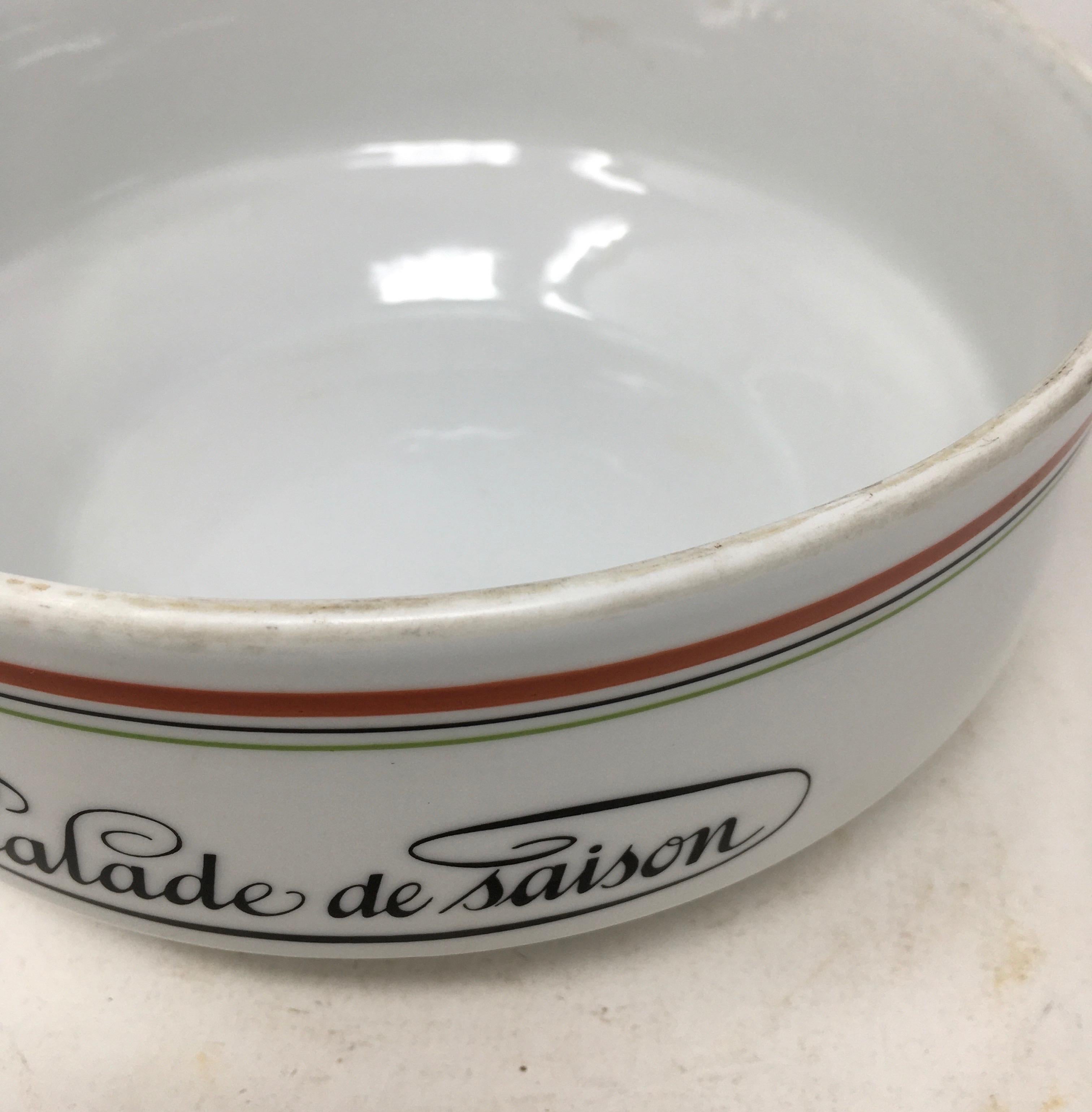 This is a lovely salad bowl created by Jacques Lobjoy. The piece is white porcelain with a red stripe and a thinner green and black stripe on the exterior. Salade de Saison is written on the front of the piece. The piece is marked on the bottom and