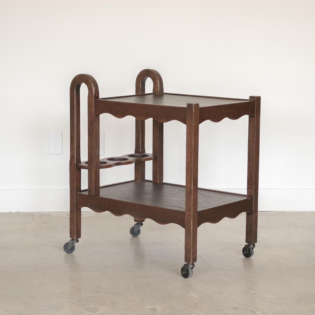Unique 1940s dark oak bar cart from France. Rectangular cart with two tiers, two curved wood handles, and 3 bottle compartments. Stunning carved wood scalloped detail on shelf edges. New metal casters. Beautiful and functional statement piece.