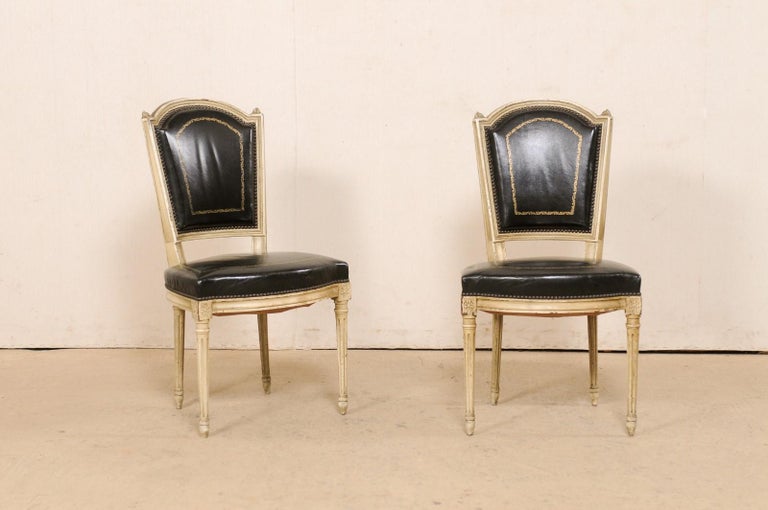 A French set of four side chairs with leather backs and seats. This vintage set of chairs from France have been carved in the typical manner of the Louis XVI period, with subtle arched top rails, finials at shoulders, floral rosettes adorning their