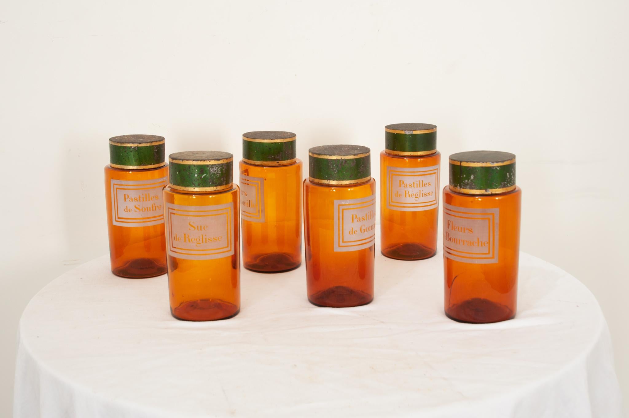 Six amber glass apothecary jars with their original green and gold tin covers from France. Stenciled, painted labels on each jar indicate their would-be contents. Three jars are complete with fitted cork tops that seal the opening under the green