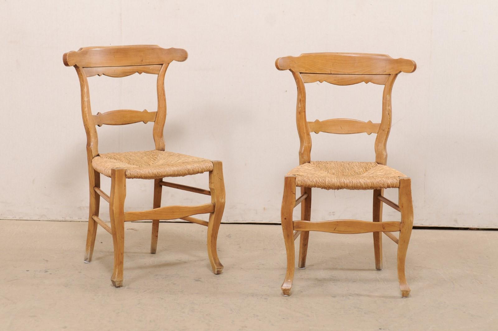 A French set of eight wooden side chairs with rush seats from the early to mid 20th century. This set of chairs from France each feature curvy carved-wood ladder-style backs, and hand-woven rush seats. These chairs are raised upon shapely carved