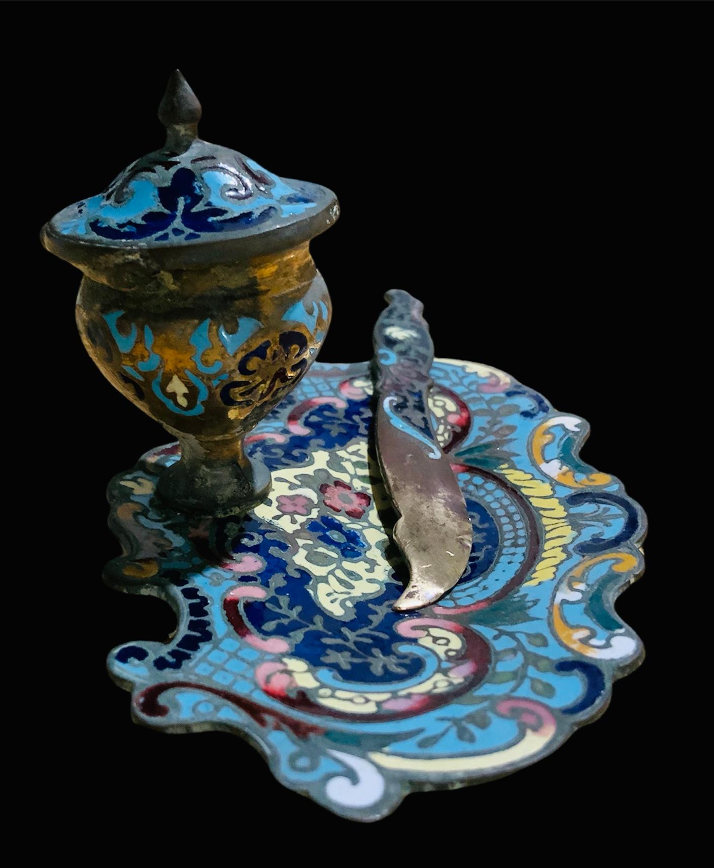 This is a small bronze champleve enamel set of an inkwell and letter opener. Both of them depicts colorful hand painted flowers, “C” scrolls and foliages. The resting plate of the inkwell depicts a serpentine border. The inkwell includes a small