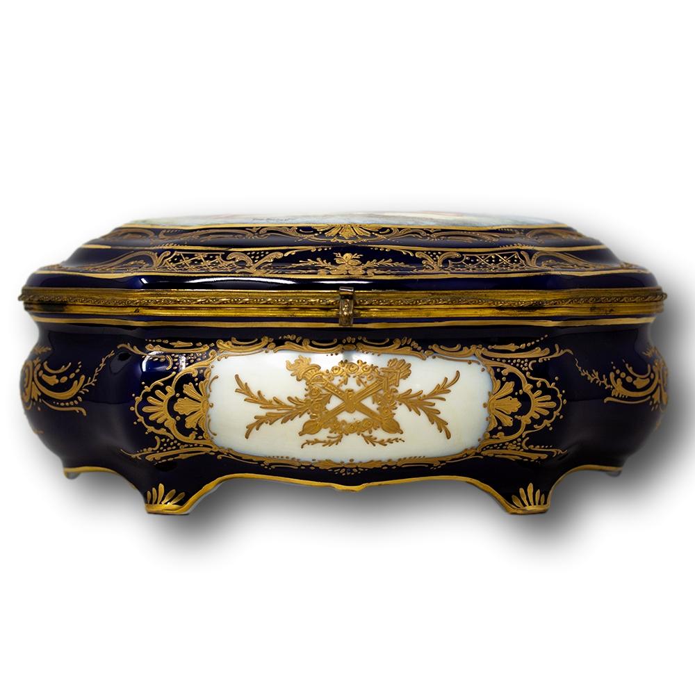 A large sized ‘Sevres‘ porcelain box. The box decorated with a base of cobalt blue with gild scrollwork decoration throughout. The lid featuring a large painted panel with a classical scene with two cupid like cherubs and a female figure after