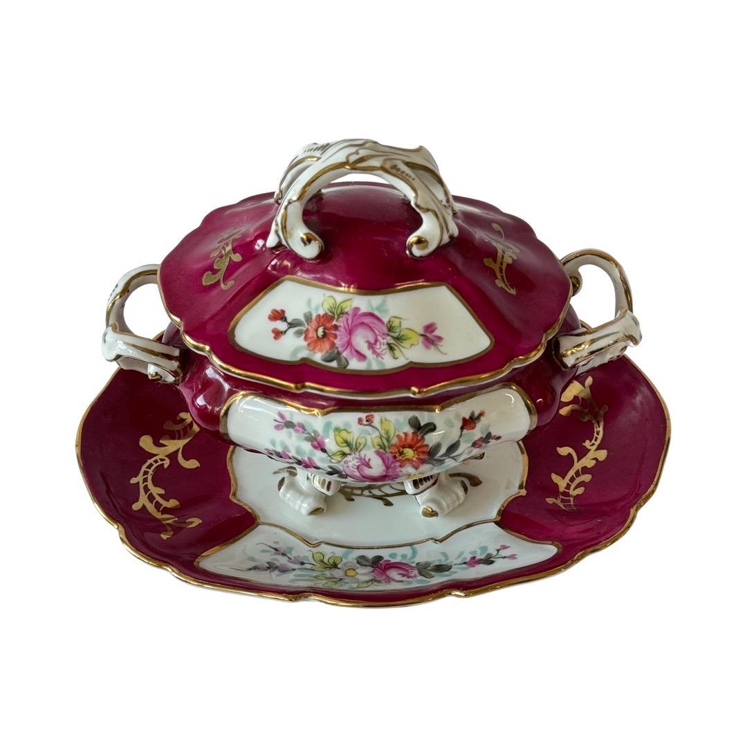 Elegante zuppiera francese in porcellana su supporto, riccamente dipinta a mano in stile Sèvres con vivaci pannelli floreali, fondo bordeaux intenso e delicati accenti dorati. Caratterizzato da manici a voluta e piedi a zampa, questo pezzo di grande