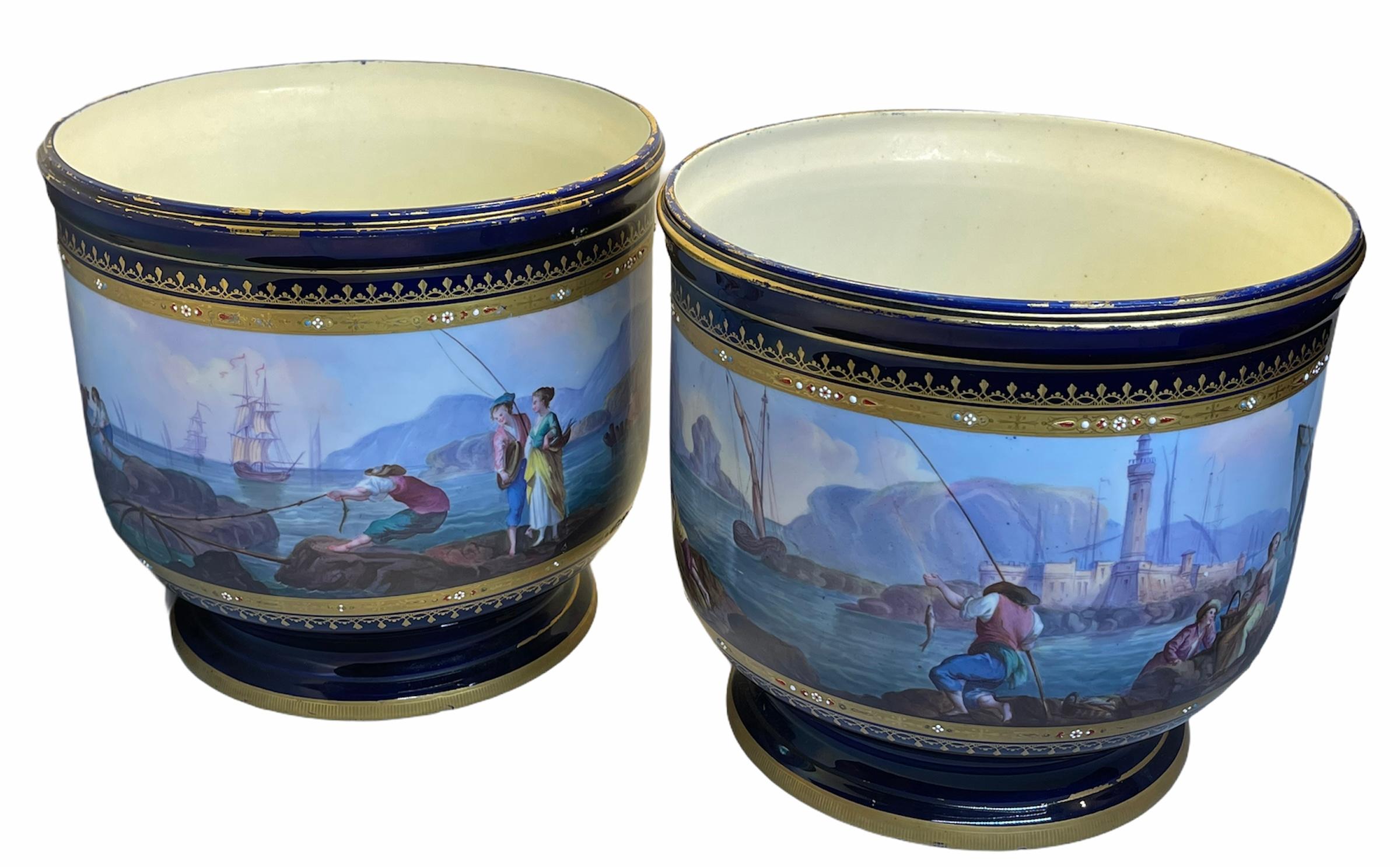 Two large gilt cobalt blue color as a background porcelain Cachepot. They are depicting a continuous hand painted scene of an 18th century coastal landscape with fishermen and ships. Embellished in the top and bottom with a golden ribbon of flowers