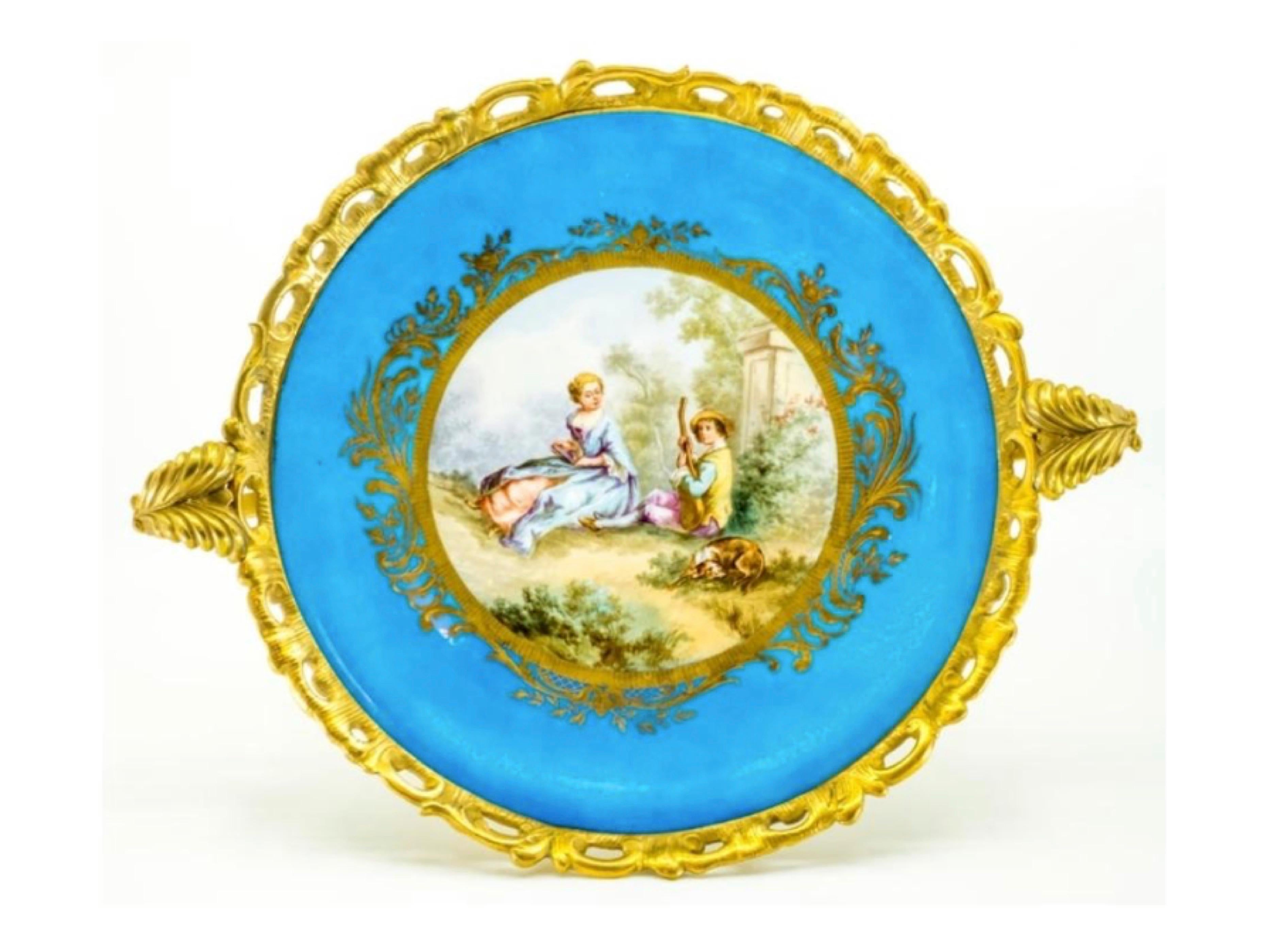 This French Sèvres-style porcelain plate from the 19th century is set in a gilt bronze mount, ormolu that features a Rococo base, rim and two handles with stylized foliage and C-scrolls.
The center of the porcelain plate displays a pastoral scene