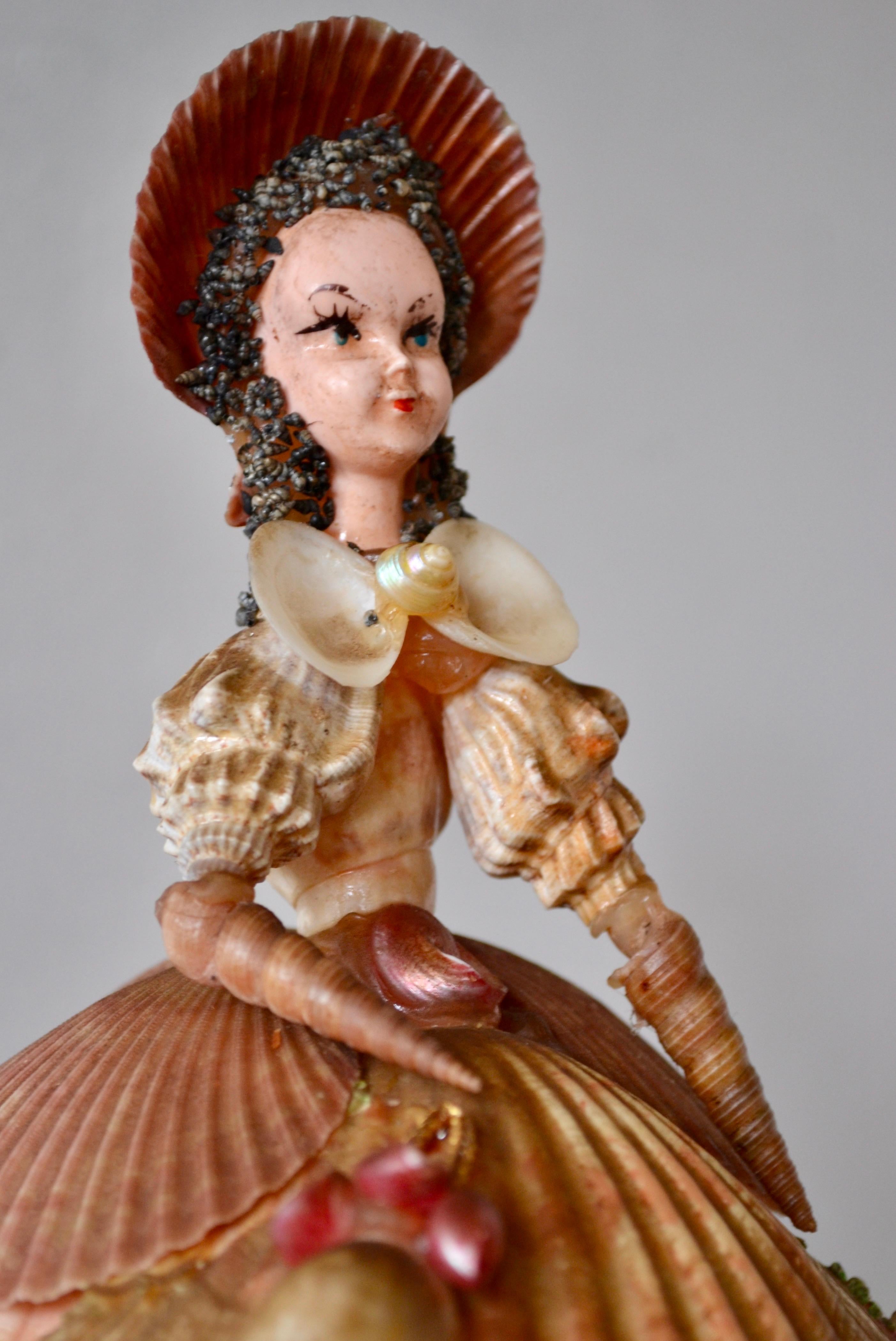 A rare and poetic decorative object, this figurine represents a lady in romantic 19th-century costume. Entirely handcrafted, the skirt is composed of scallop shells, beautifully arranged and embellished with a garland of delicate flowers. The finely