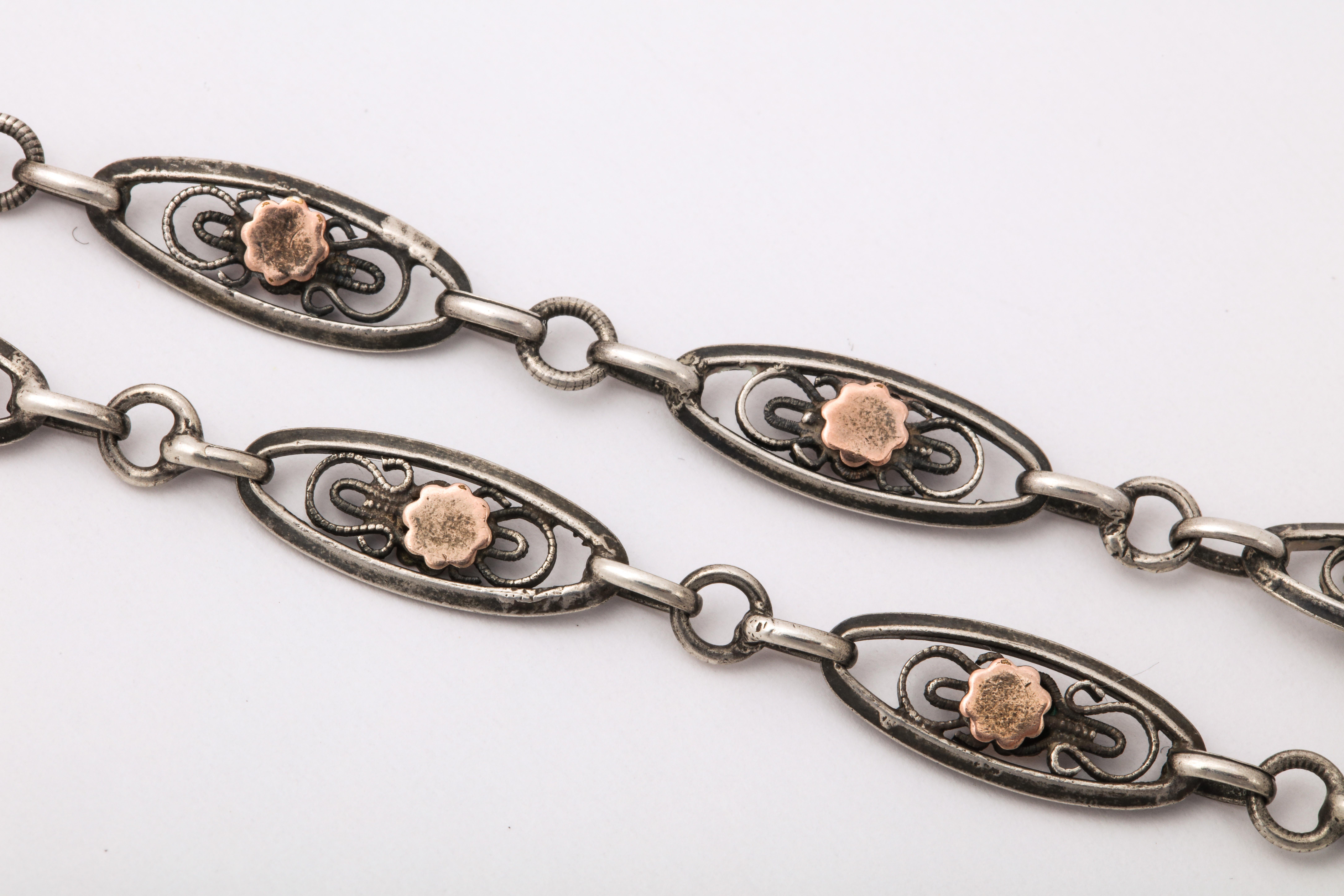 Una catena francese del 1870 brilla con un fiore astratto in oro al centro di un simbolo dell'infinito all'interno di una maglia ovale in argento. La catena è una di quelle che ami toccare. È setoso e liscio. È eccezionale su ogni colore di tessuto,