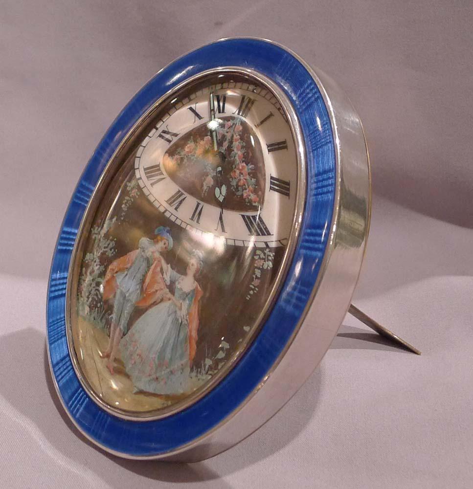 French silver and guilloche enamel timepiece with hand painted picture.
Of unusual construction with oval case decorated to the rim with a one inch border in deep blue guilloche enamel. The face with triangular or fan shaped chapters below the 12