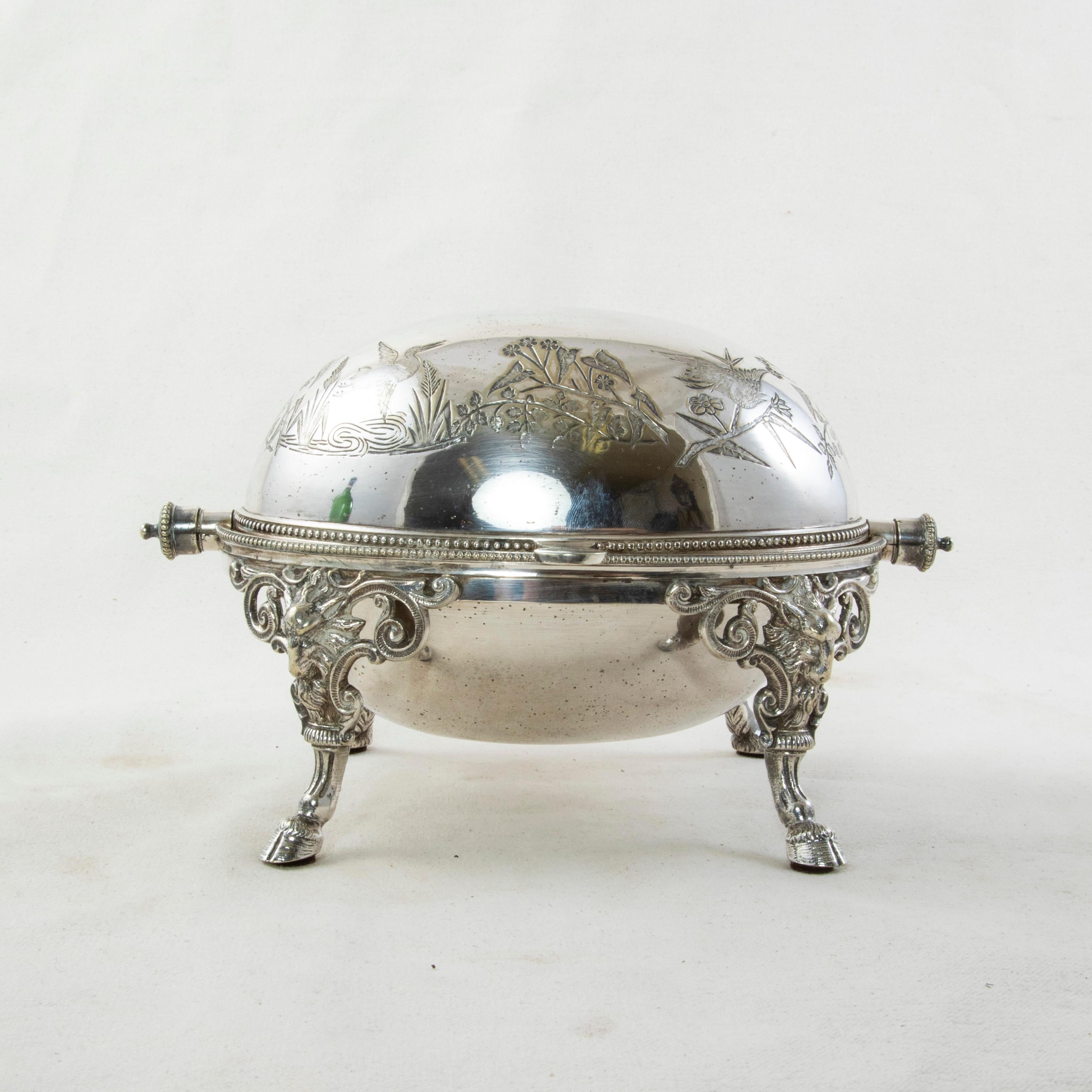 This French silver plate domed serving piece from the turn of the twentieth century features a swiveling lid engraved with egrets and a floral motif. The domed bowl rests on curved legs detailed with a scrolling pattern, rams heads, and hooved feet.