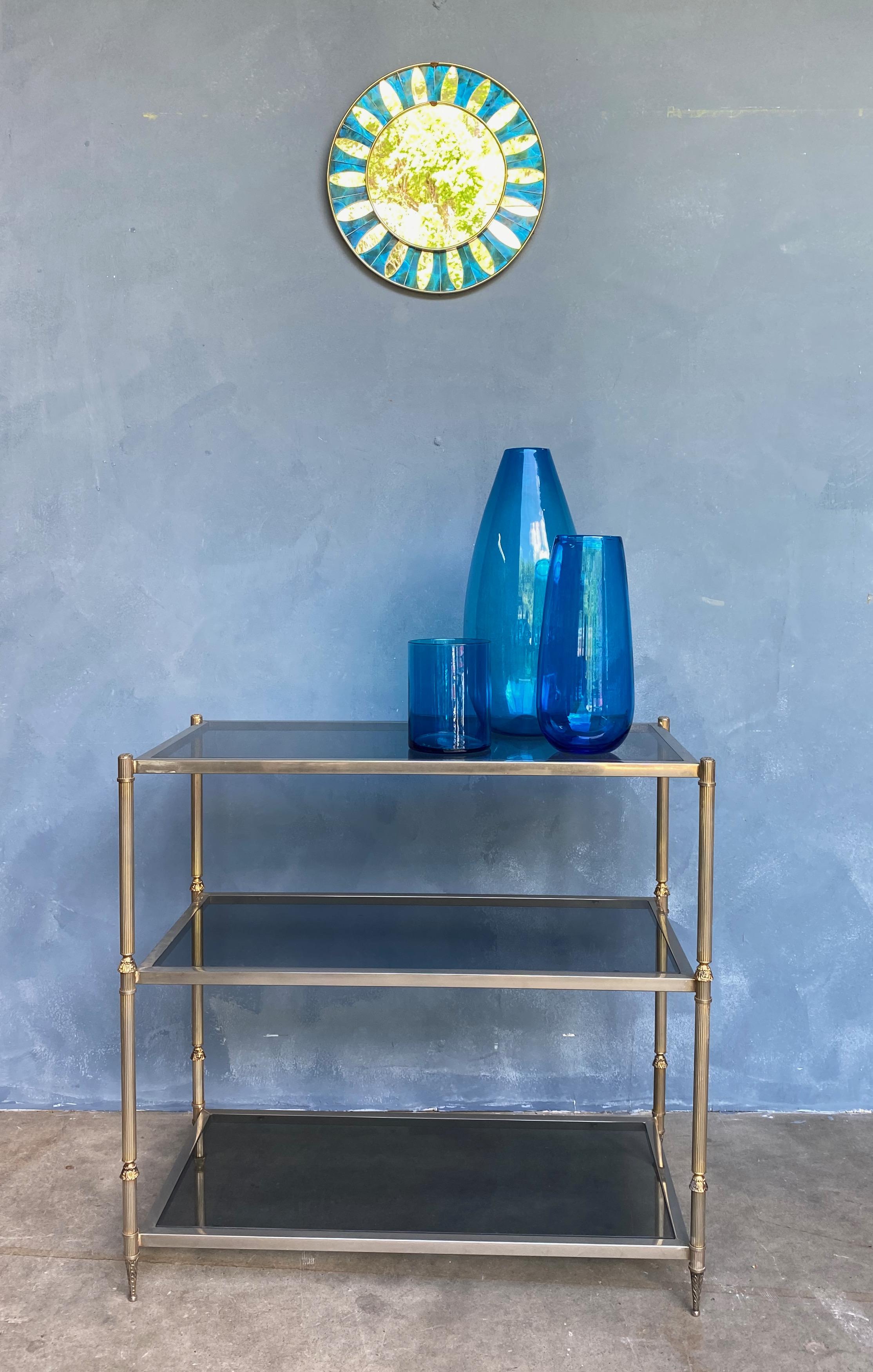 This handsome French brass and glass 3 tiered console hails from the 1970’s. It features three smoked glass shelves enclosed in a patinated silvered brass frame, with fluted brass legs and classical motifs providing an elegant touch. The vintage