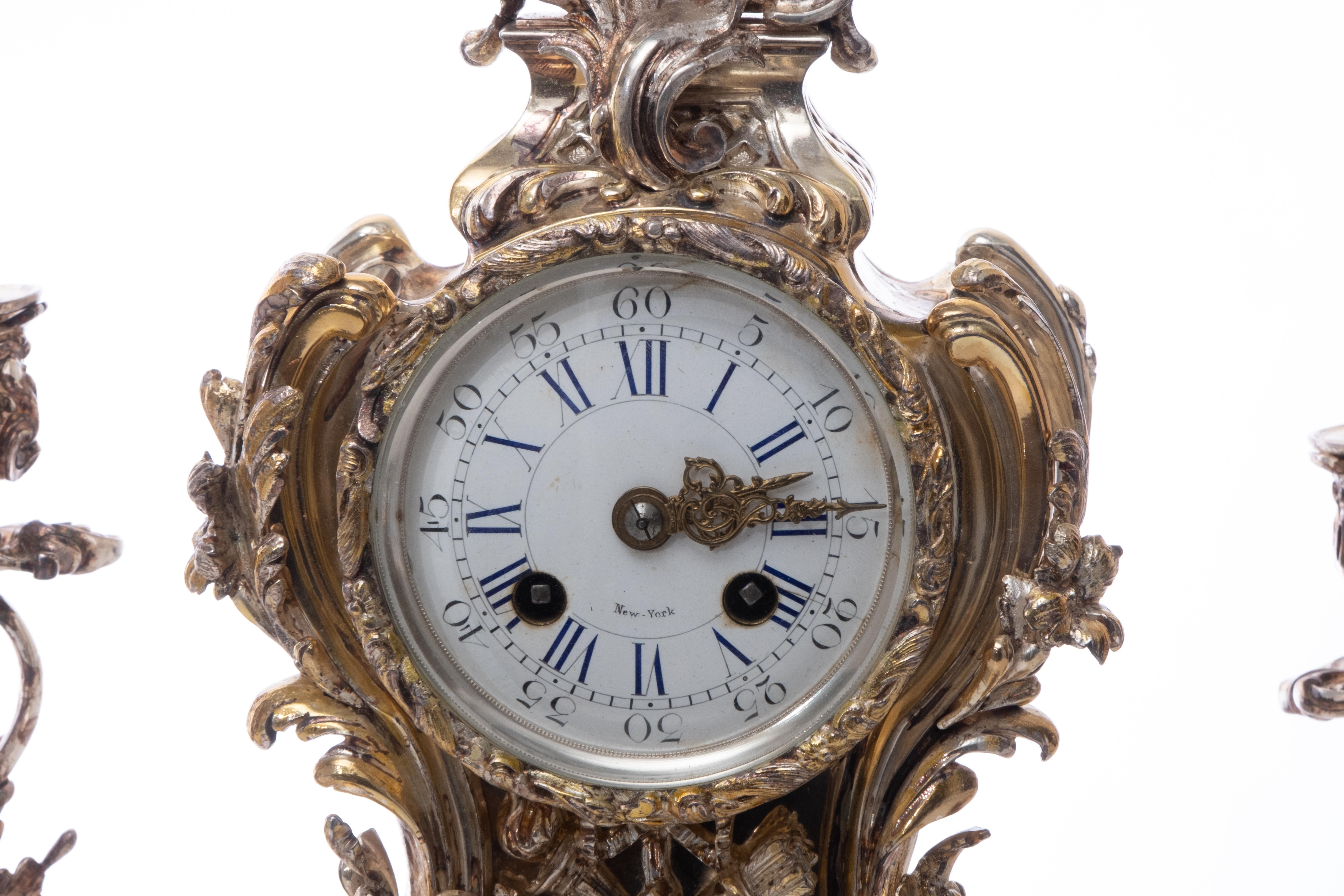 A French Louis XIV three (3) piece silver over bronze mantel clock and garnitures set. A clock in silver over bronze accompanied with a pair of candlestick garnitures. Produced in an elegant Baroque King Louis VI design. White enamel clock face with