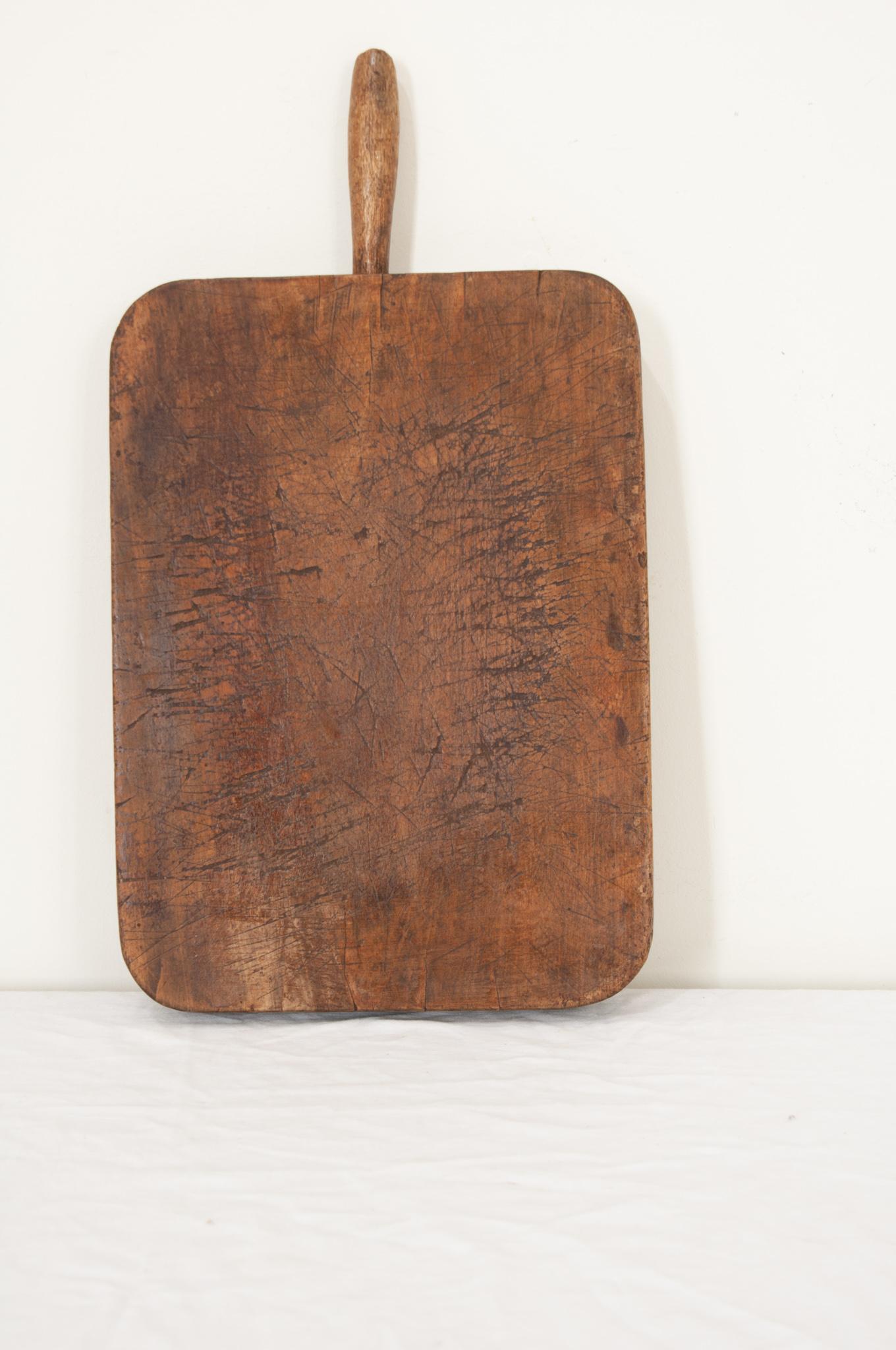 A large solid wood cutting board from France in a rectangle shape with smooth corners and a handle. The worn board is made of a single piece of wood. Knife marks and scrapes that were left by cooks, now long gone, can be seen and felt as if they