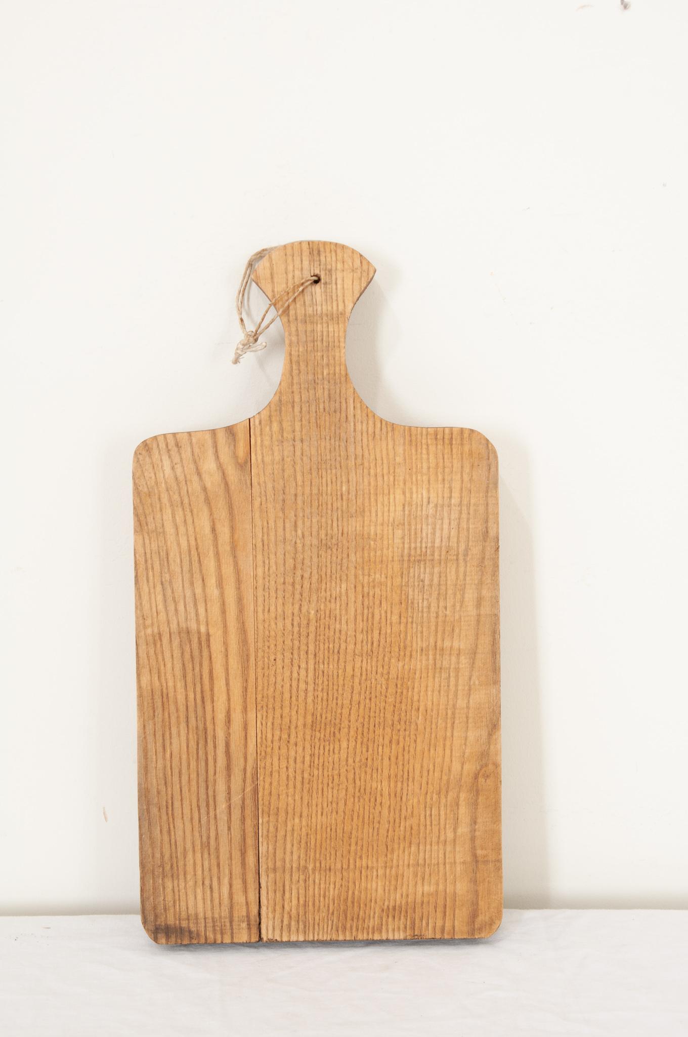 A large single plank wood cutting board from France in a paddle shape with smooth corners and a handle with option to hang on the wall. The wood has a nice patina and wonderful striations in the wood grain. Knife marks and scrapes that were left by