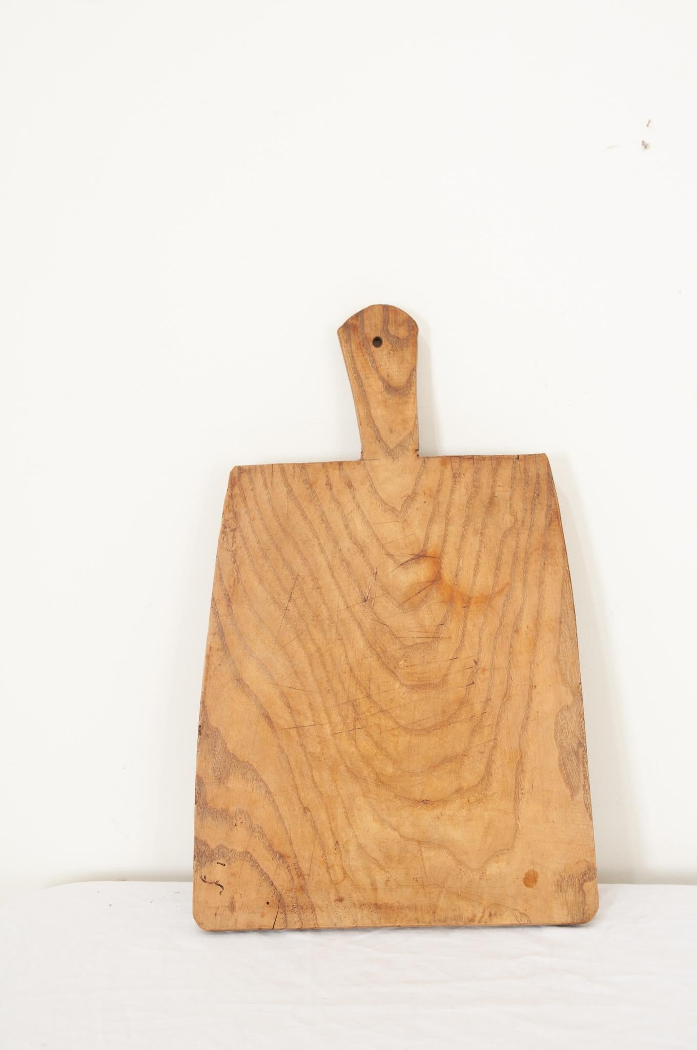 A large single plank wood cutting board from France in a paddle shape with smooth corners and a handle. Knife marks and scrapes that were left by cooks, now long gone, can be seen and felt as if they were made yesterday. A nice antique, full of