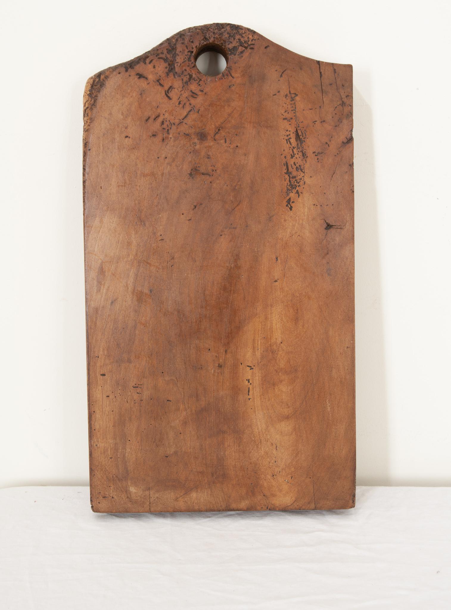 A very large 2 ⅜” thick single plank wood cutting board from France with shaped smooth corners and a circular cut-out that creates a short handle. Knife marks and scrapes that were left by cooks, now long gone, can be seen and felt as if they were