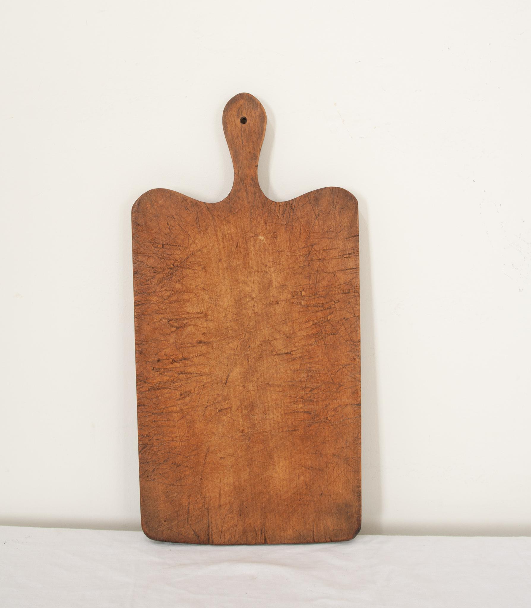 A solid wood cutting board from France in a rectangle shape with smooth corners and a handle. The worn board is made of a single piece of wood. Knife marks and scrapes that were left by cooks, now long gone, can be seen and felt as if they were made